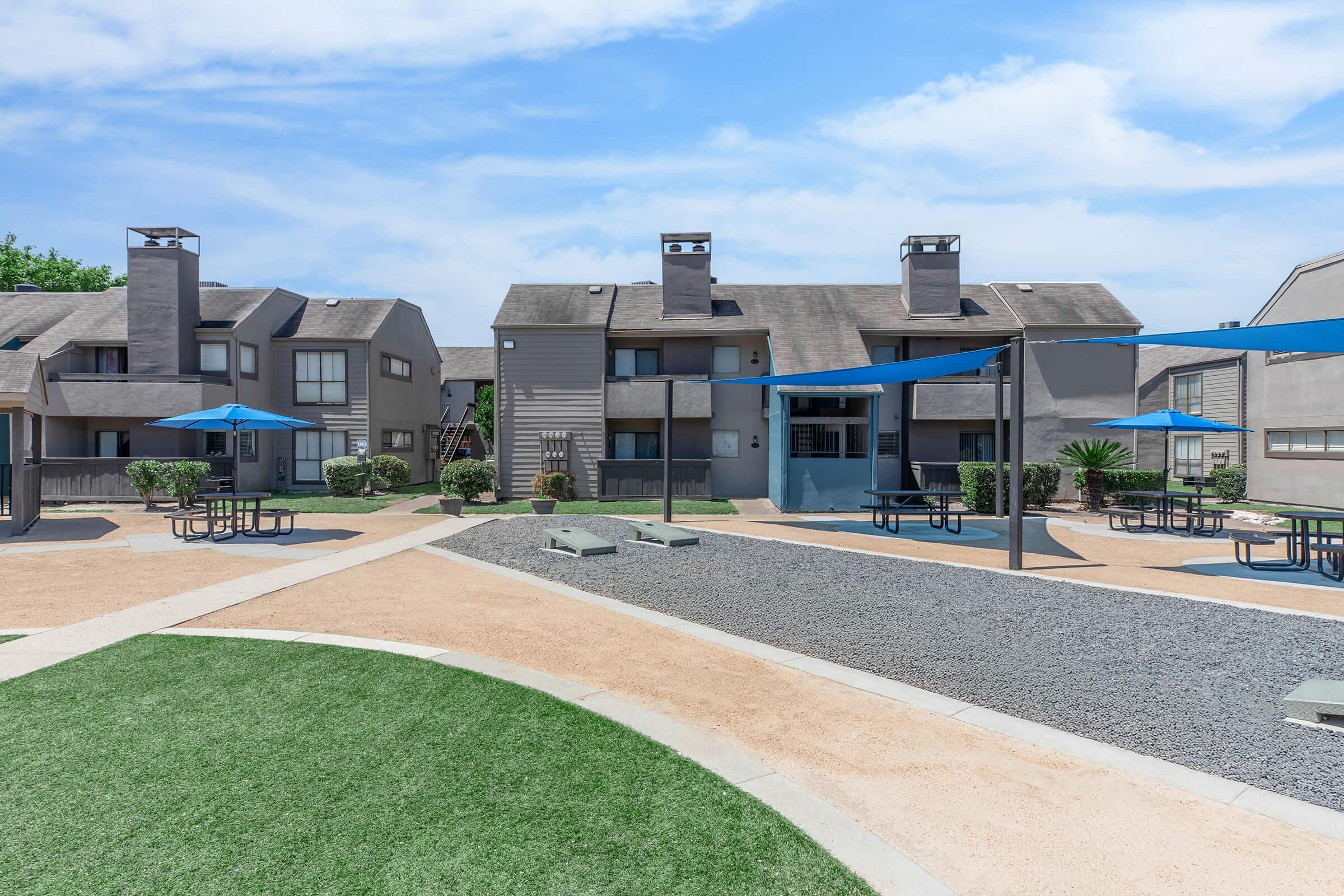View of a residential complex featuring two-story buildings, equipped with blue umbrellas and picnic tables in a landscaped outdoor area. The scene includes grassy patches, pathways, and decorative gravel. The sky is clear with a few clouds, creating a vibrant and inviting atmosphere.