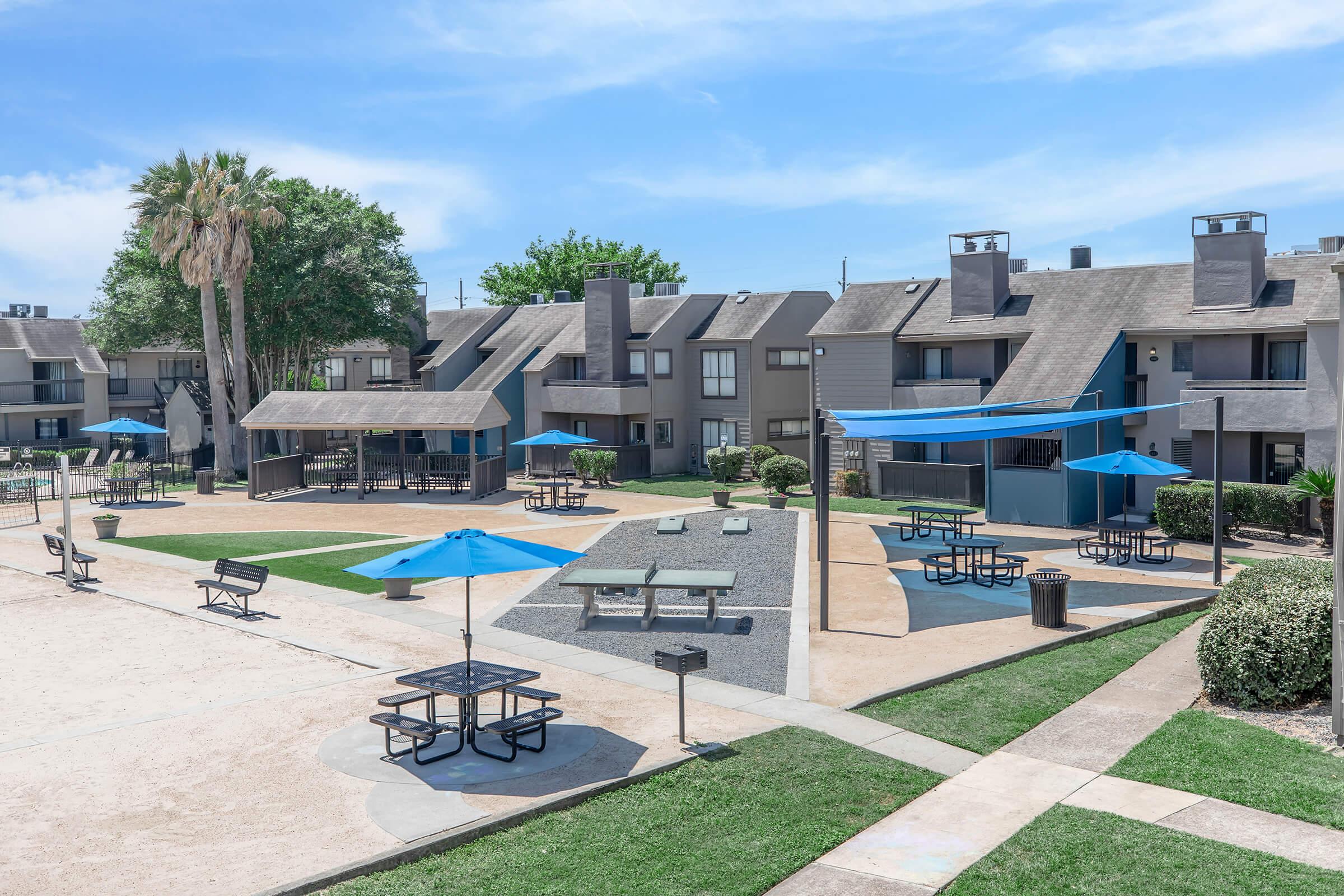 A residential complex featuring several two-story buildings surrounded by green grass. In the foreground, picnic tables with blue umbrellas are arranged in a courtyard area, which includes pathways, a shaded pavilion, and landscaped bushes. The sky is clear with a few clouds.