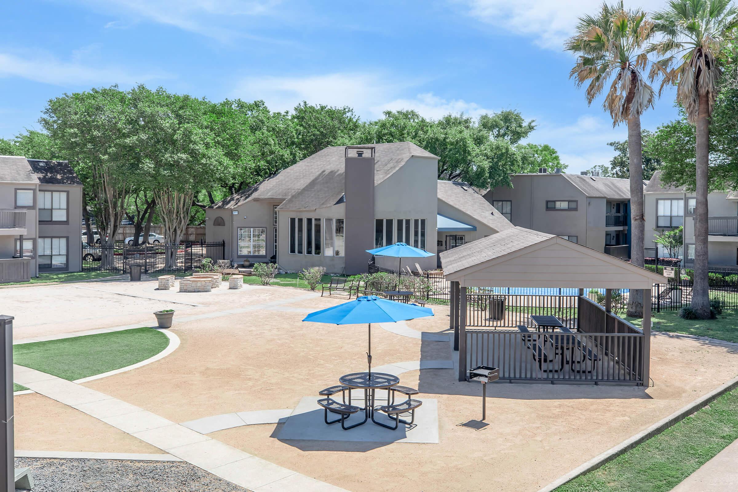 A landscaped apartment complex featuring a grassy area with picnic tables and blue umbrellas, surrounded by trees and residential buildings. There are designated spots for grilling and a communal gathering area, all under a clear blue sky.