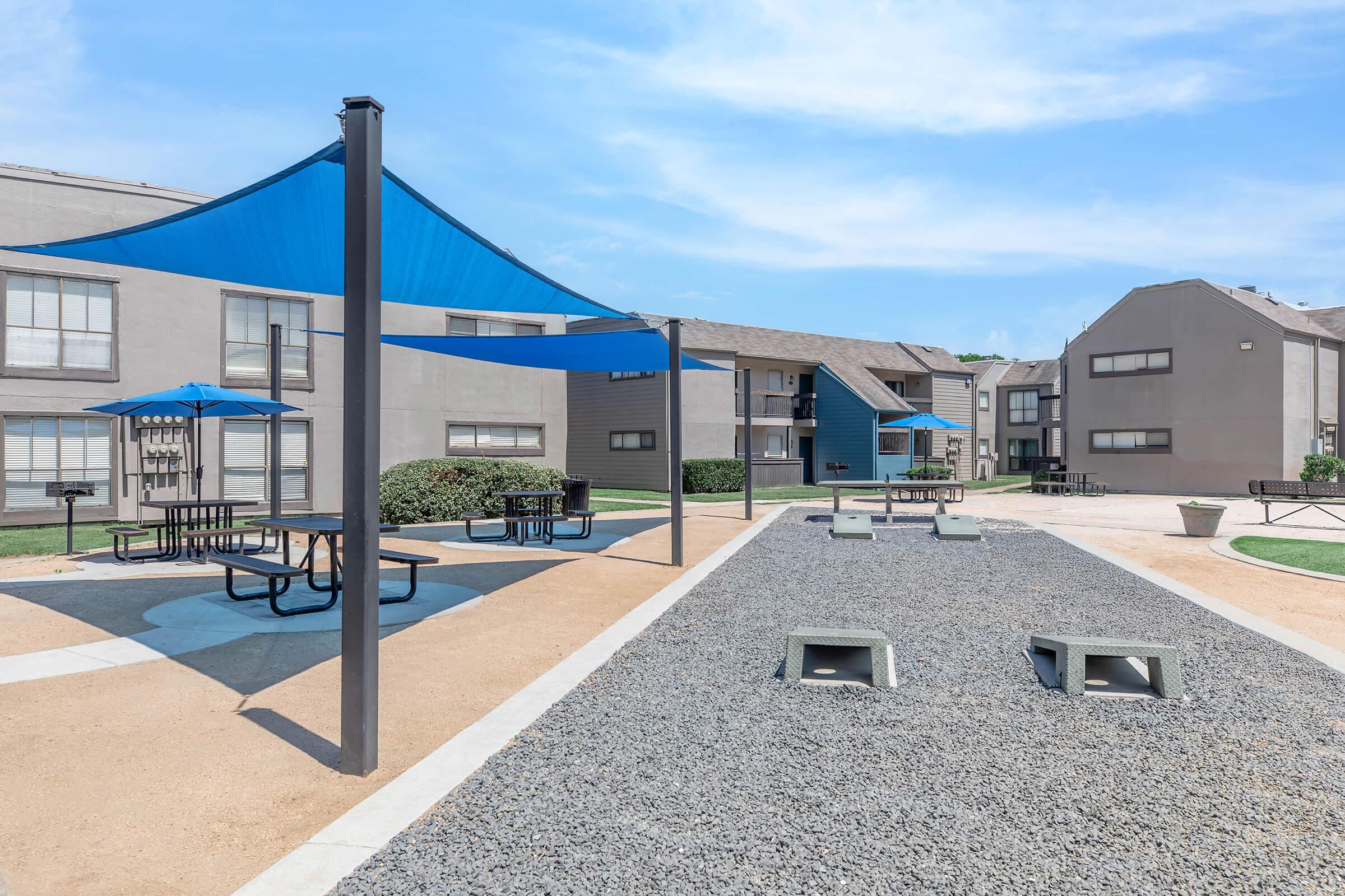 Outdoor recreational area with shaded seating under blue canopies, picnic tables, and a gravel path. Surrounding buildings are gray with multiple windows, and there is a grassy area in the background. The sky is clear, showcasing a bright, sunny day.