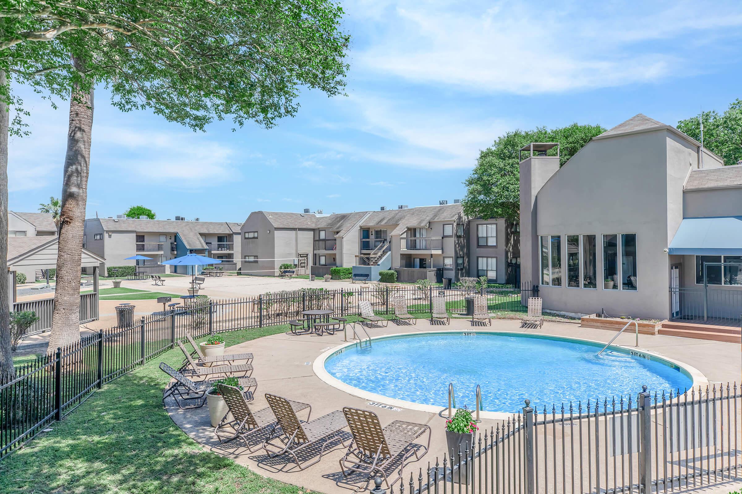 A view of an outdoor pool surrounded by lounge chairs, with a landscaped area and several apartment buildings in the background. A tree provides shade near the pool, and the sky is clear and blue. The setting appears to be a residential community with well-maintained grounds.