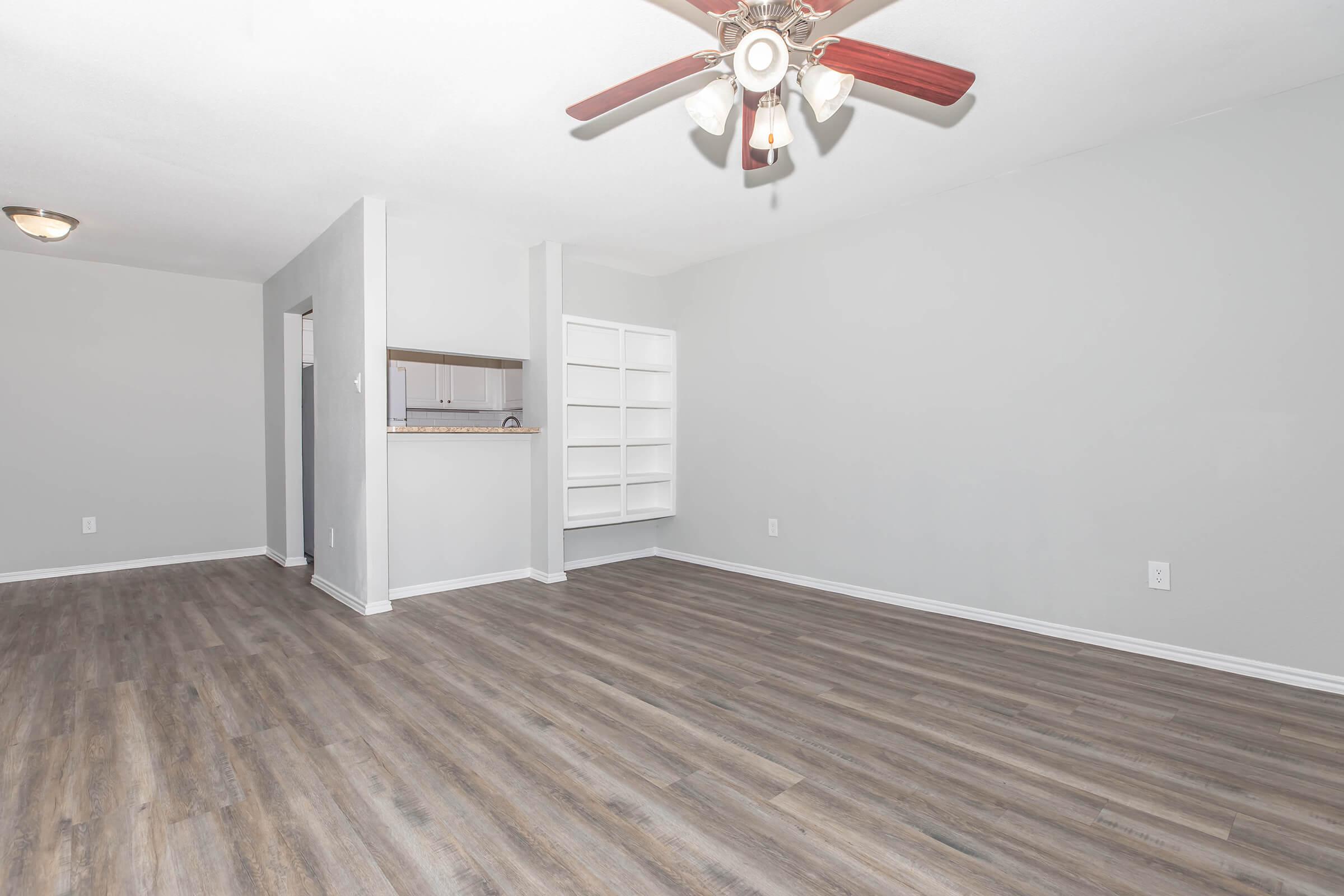 A spacious, empty living room featuring light gray walls and wooden flooring. There is a ceiling fan in the center and a small open shelving unit on one side. In the background, an entryway leads to a kitchen area. The space is bright and unobstructed, ready for decoration.