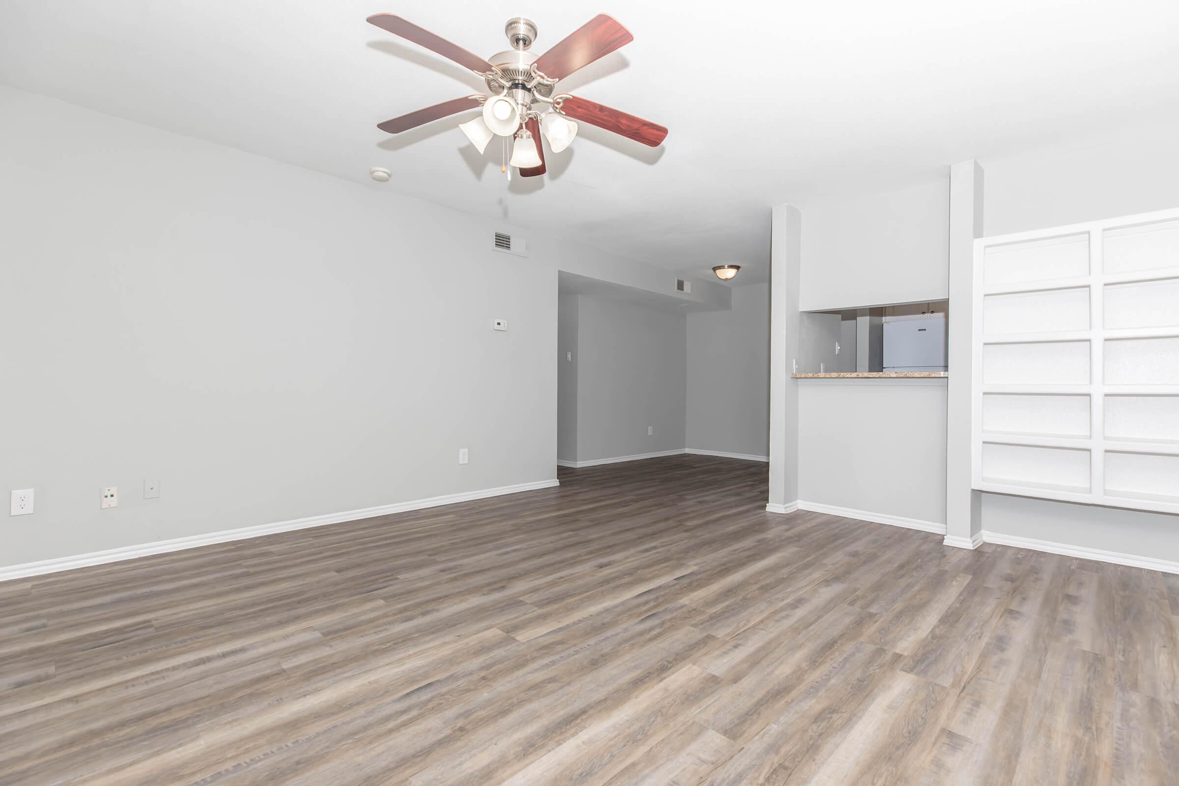 A spacious, empty living room featuring gray walls, a ceiling fan with wooden blades, and laminate flooring. There is a partial wall with a small opening that leads to a kitchen area, and a built-in shelf against one wall. The room is well-lit with natural light and has a clean, modern aesthetic.