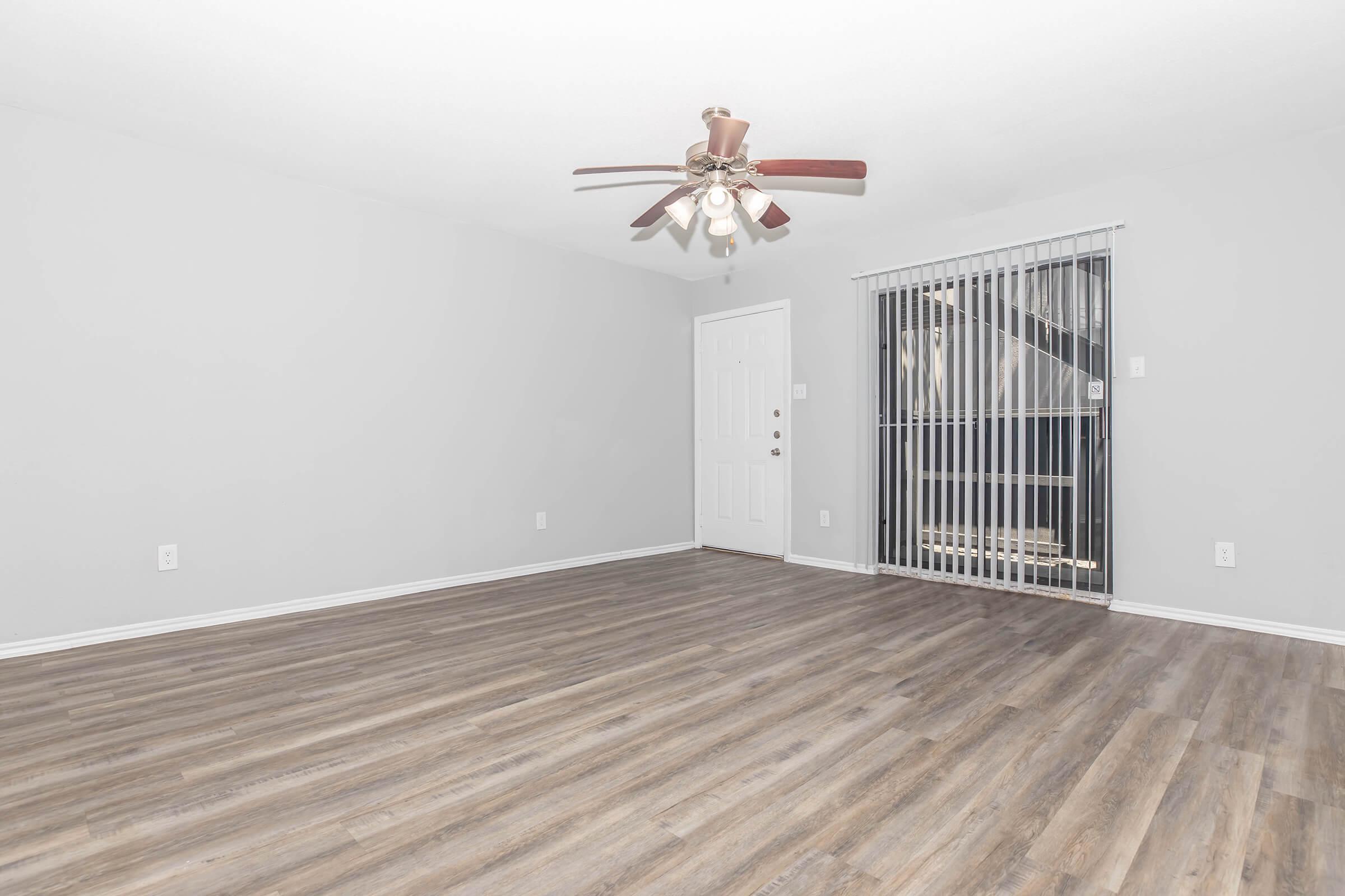A spacious, empty room with light gray walls and new wood-style flooring. A ceiling fan with wooden blades hangs above. On one side, there is a door leading outside and vertical blinds covering a window. A staircase is partially visible in the background, emphasizing the open layout of the space.