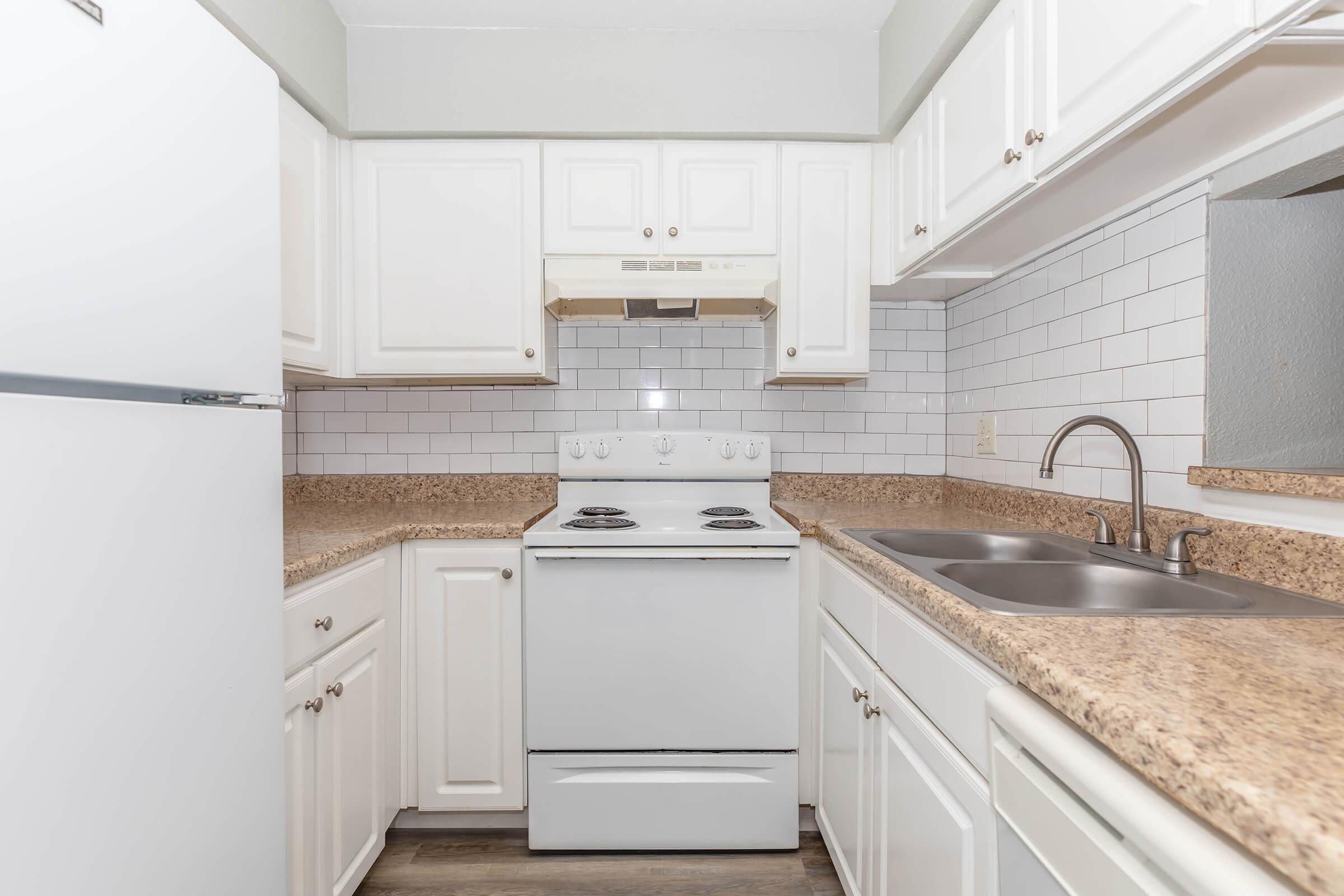 A modern kitchen featuring white cabinetry, a light-colored countertop, a stove with an oven, a hood above, and a dual sink. The backsplash is tiled in white, and there is a refrigerator on the left side, creating a clean and bright cooking space.