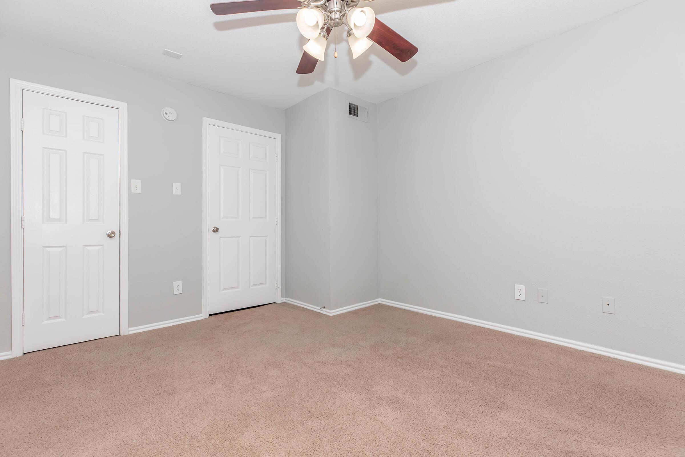 Empty room with light gray walls and beige carpet. A ceiling fan with four blades is overhead. Two closed white doors are visible on the left side, and a light switch appears on the wall. The room has a clean, minimalist appearance with no furniture or decorations.