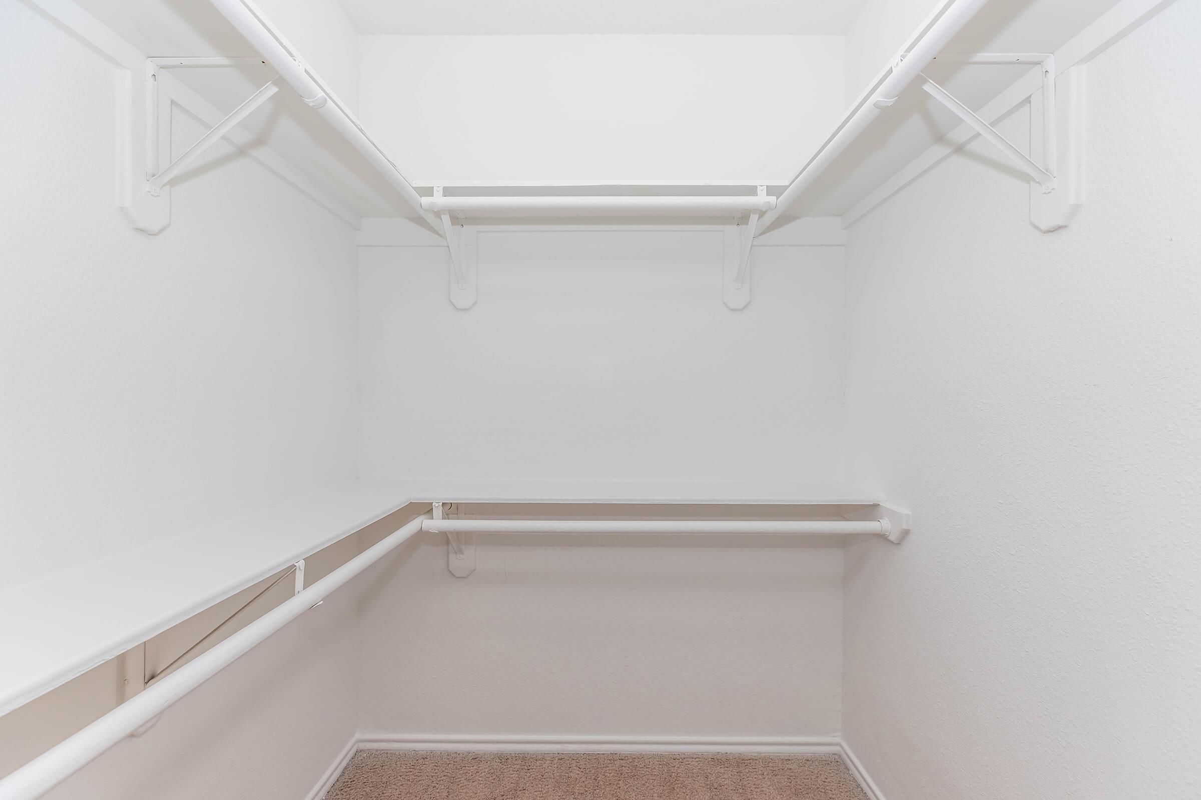 Empty walk-in closet with white walls and shelving. The closet features two horizontal shelves and multiple hanging rods, with a carpeted floor. The overall space is well-lit and minimalist, providing a clean and organized appearance.