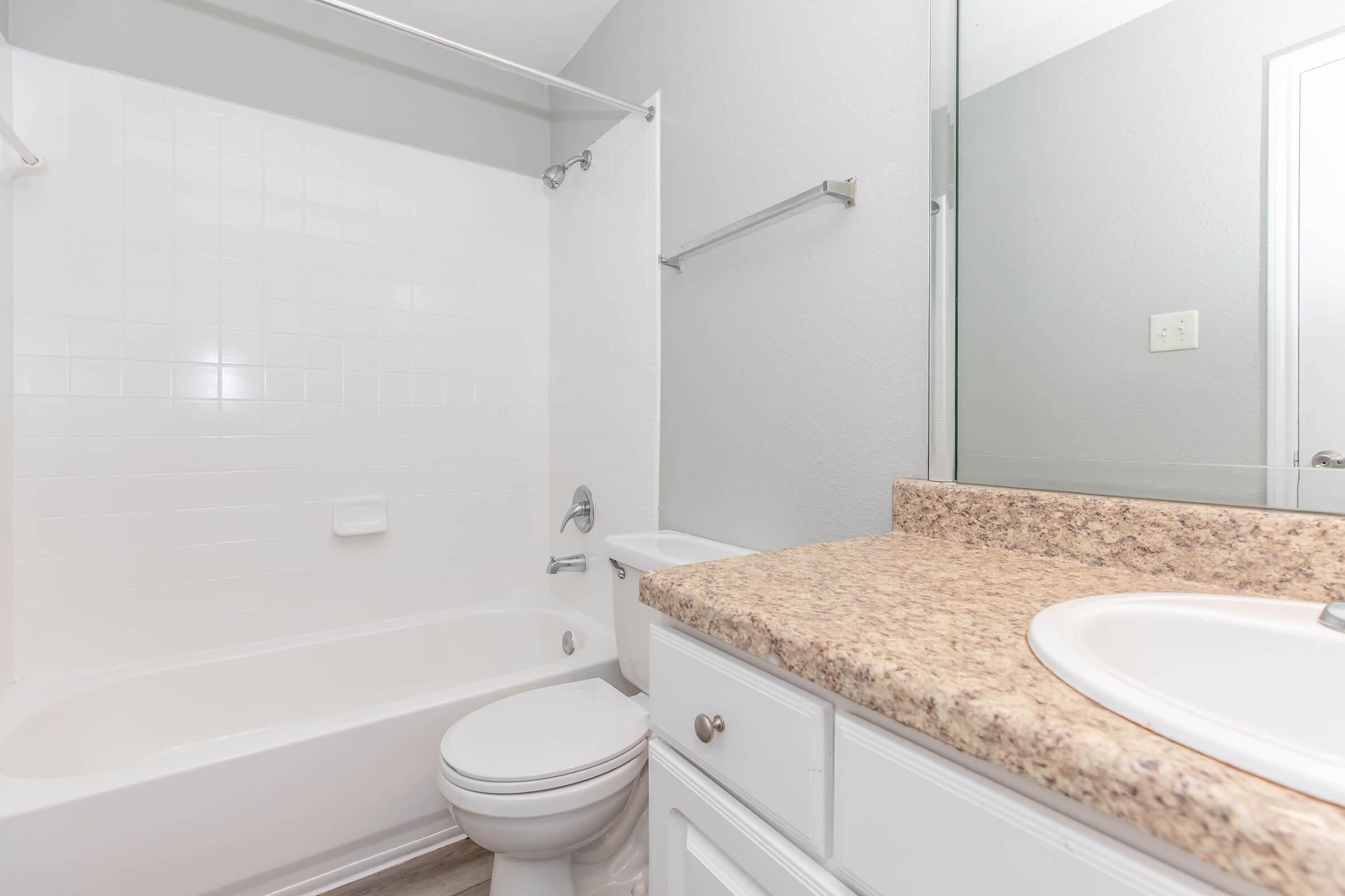 A clean and modern bathroom featuring a white bathtub with a shower, a granite countertop with a sink, and a toilet. The walls are light gray, and a large mirror is above the sink, enhancing the spacious feel of the room.
