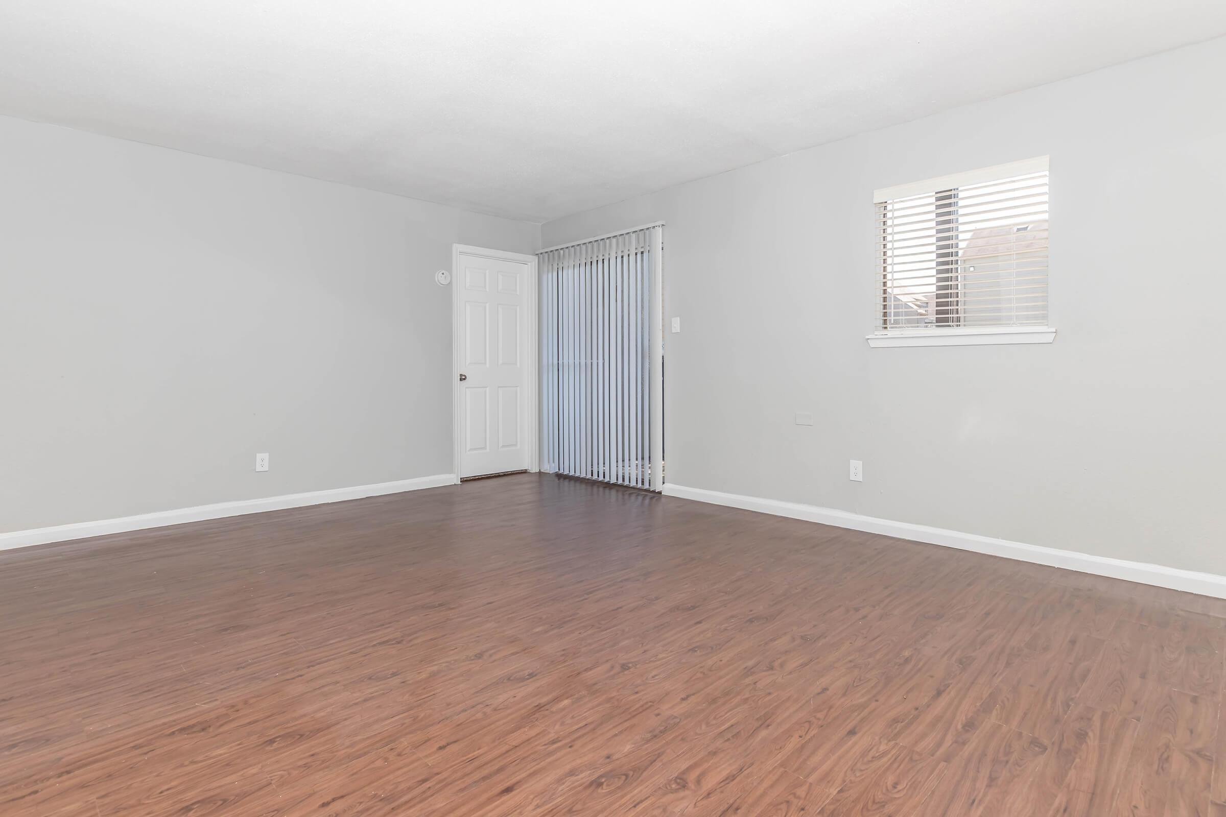 A spacious, empty room with gray walls and a glossy wooden floor. There is a door on the left, and a window with blinds on the right, allowing natural light to enter. The overall ambiance is bright and minimalistic, suggesting a versatile space suitable for various purposes.