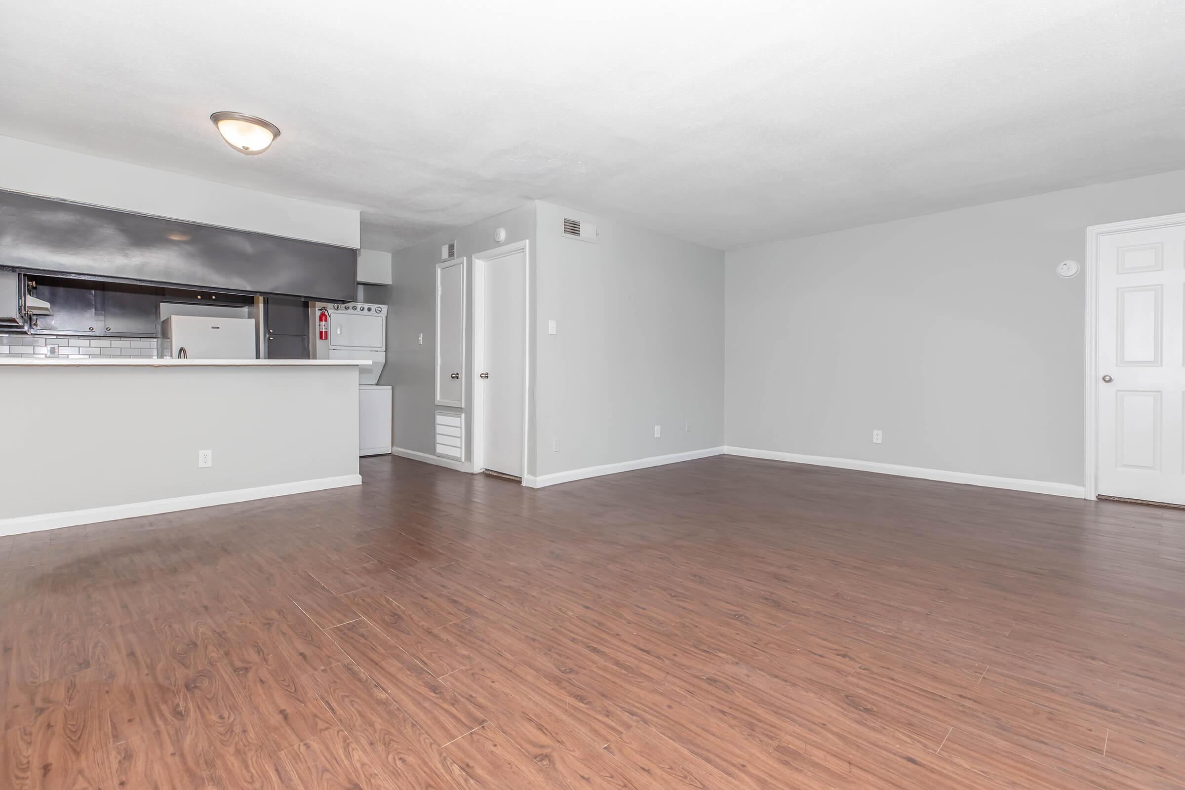A spacious, vacant living area with light gray walls and medium wood laminate flooring. To the left, a kitchen area is visible, featuring dark cabinets and a white refrigerator. The room has ample natural light and a modern feel, with one door leading to another room and a second door possibly to a closet.