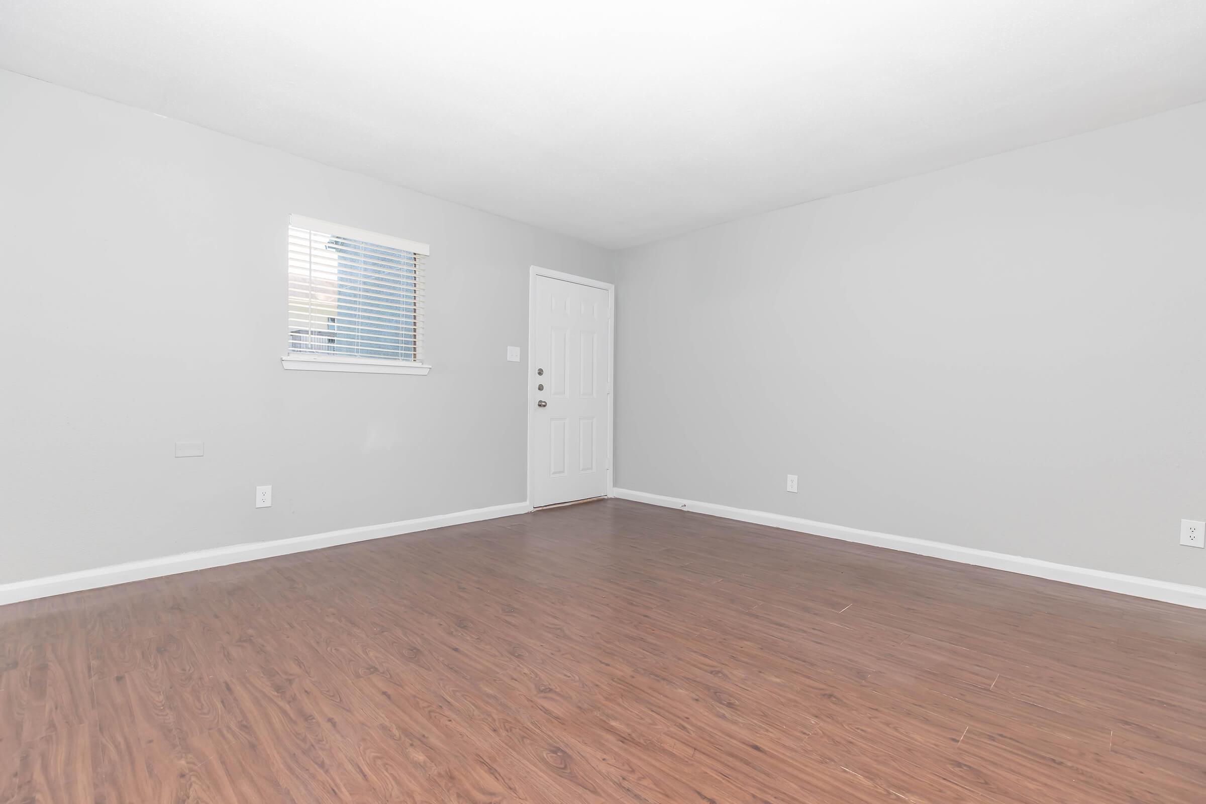 An empty room with light gray walls, a wooden floor, a small window with blinds, and a white door. There are no furnishings or decorations, creating a spacious and minimalist appearance.