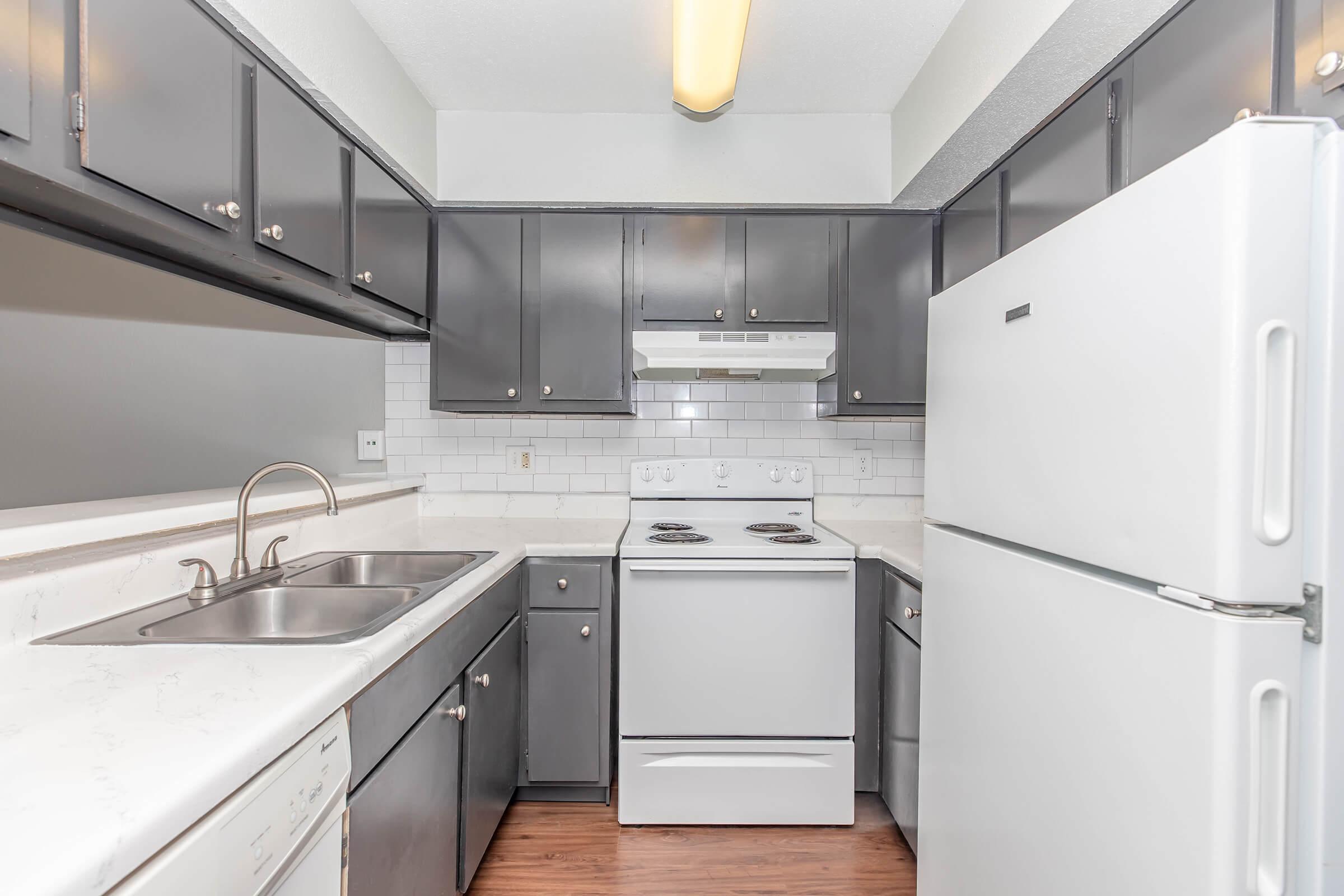 A modern kitchen featuring gray cabinets, a white stove, and oven combination, a double sink with a polished chrome faucet, and a refrigerator. The countertops are light-colored, and the backsplash consists of white subway tiles. There is a ceiling light fixture providing illumination.
