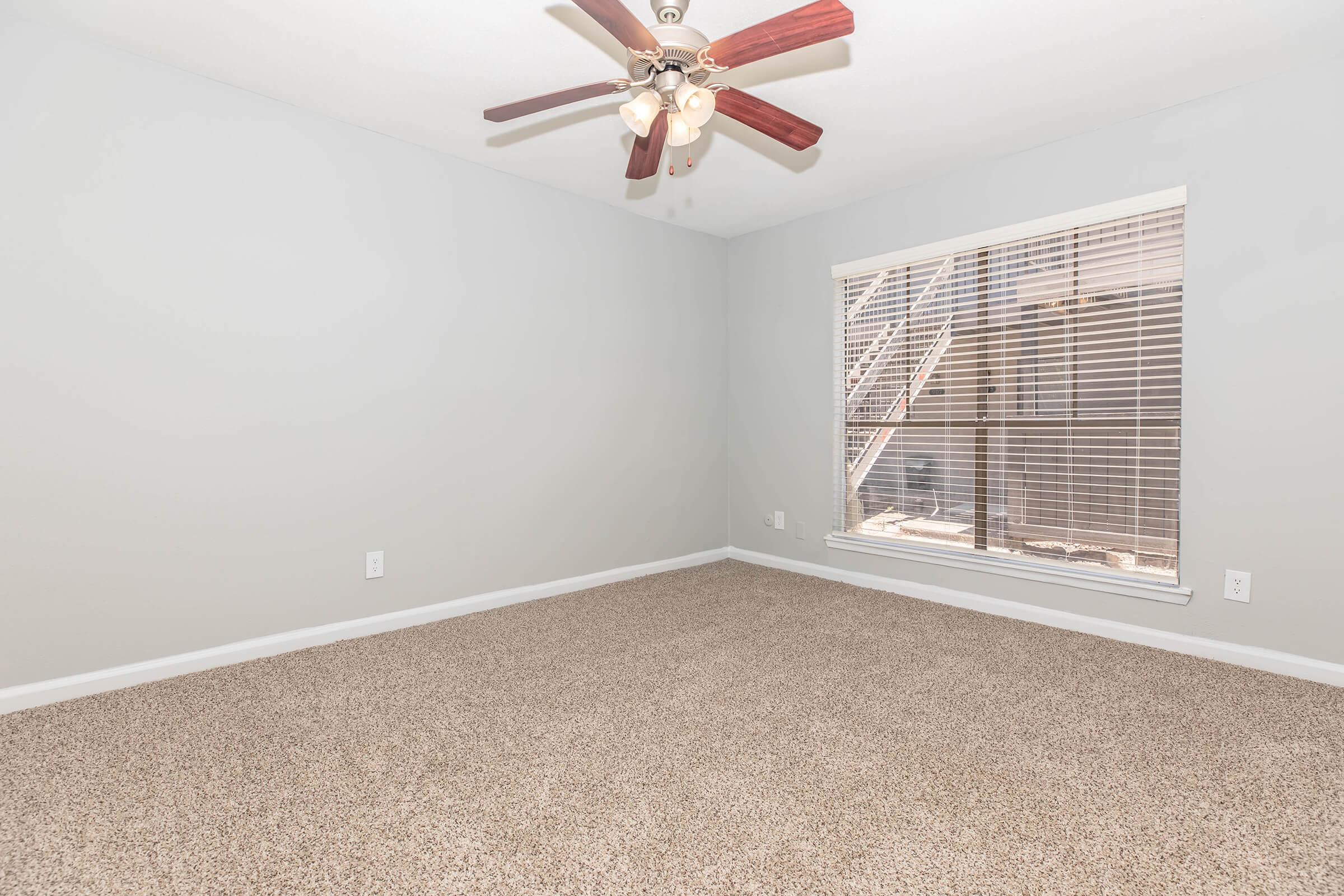 Empty room with light gray walls and a ceiling fan with wooden blades. The floor is covered in plush beige carpeting. A window with blinds lets in natural light, revealing a glimpse of an exterior staircase outside.