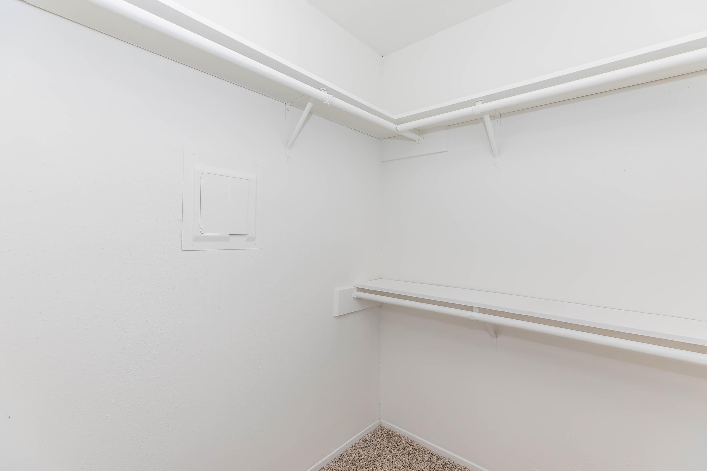 Empty closet with white walls, featuring two horizontal shelves and a small access panel on the left side. The floor is covered with a beige carpet. The space is well-lit, emphasizing its clean and minimalistic design.