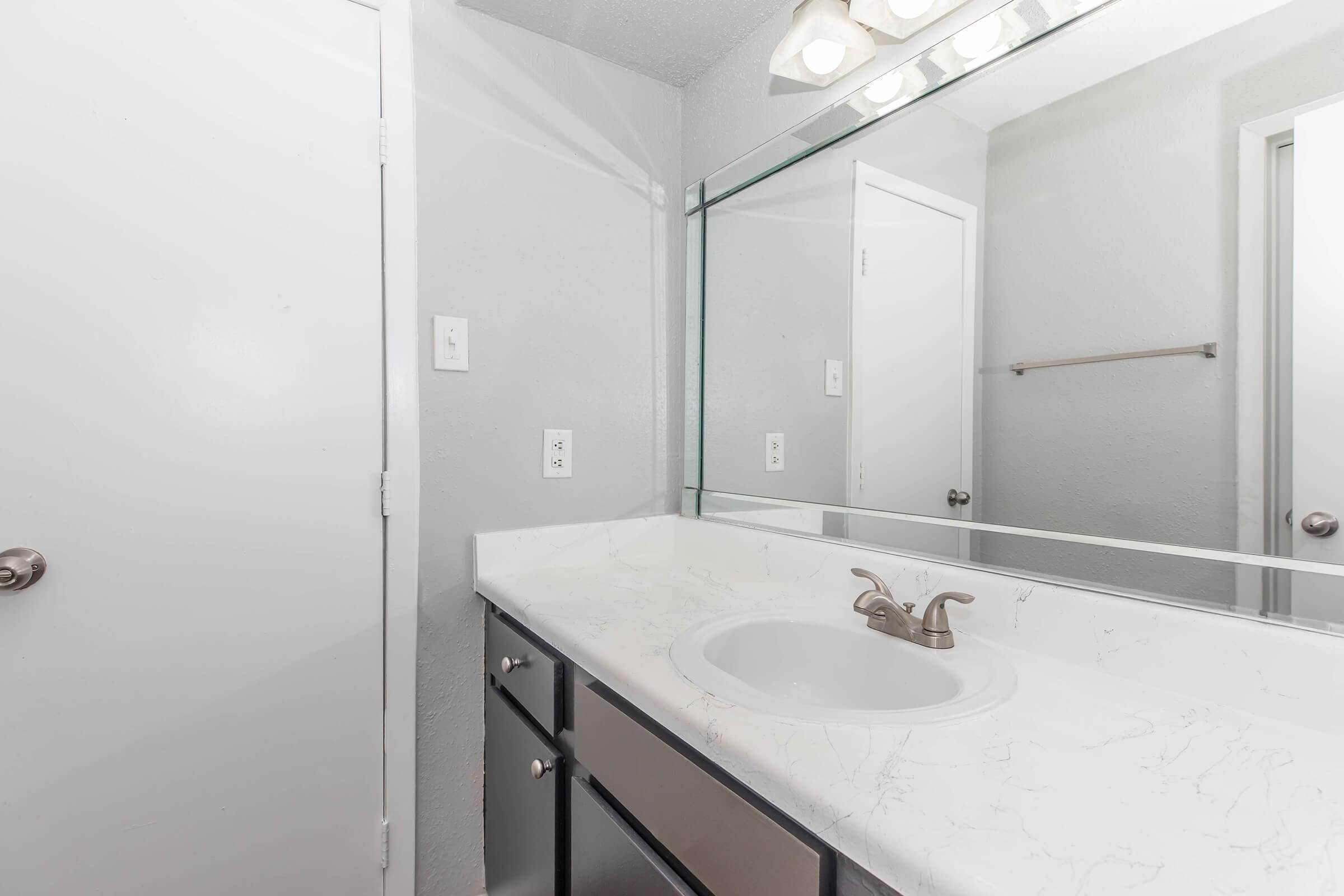 A clean and modern bathroom featuring a white countertop with a sink, a mirror above, light fixtures, and gray walls. There are doors in the background leading to other areas, and a towel bar is visible near the sink area. The overall aesthetic is minimalist and well-lit.