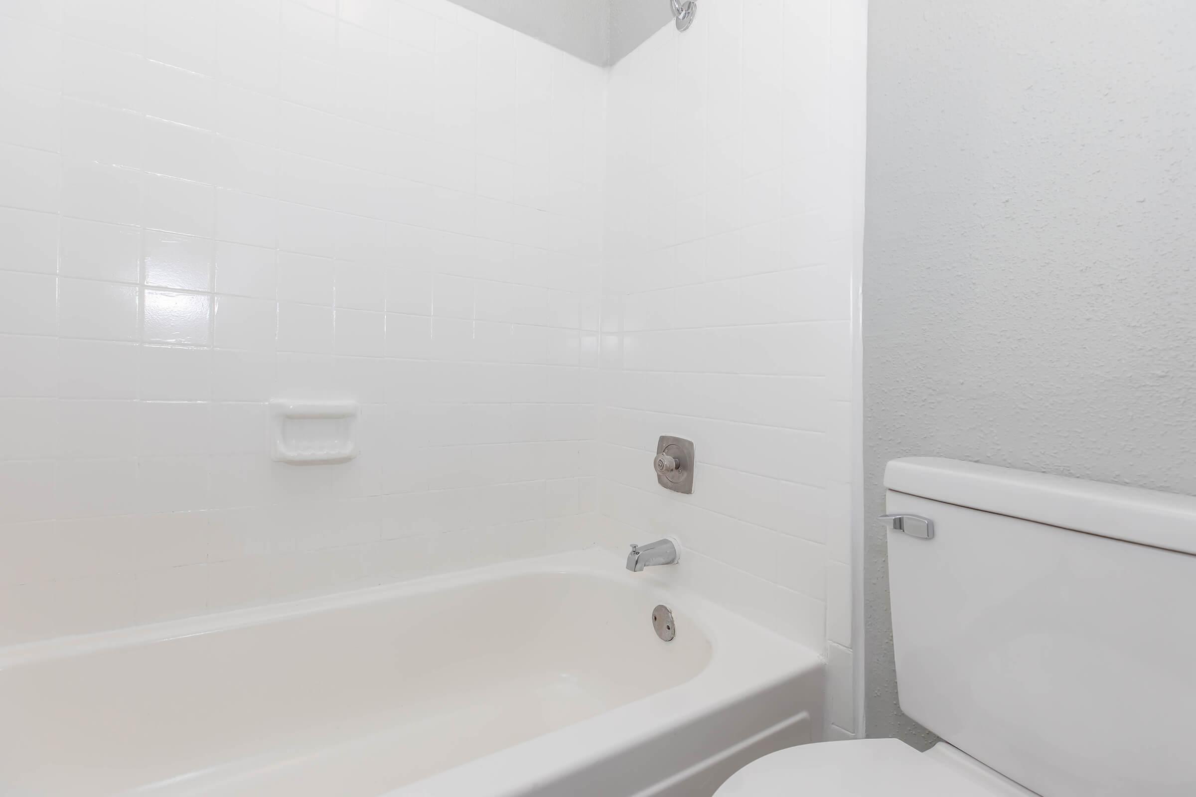 A clean, simple bathroom featuring a white bathtub, a showerhead on the wall, and a toilet. The walls are tiled in white, creating a bright and minimalist appearance.