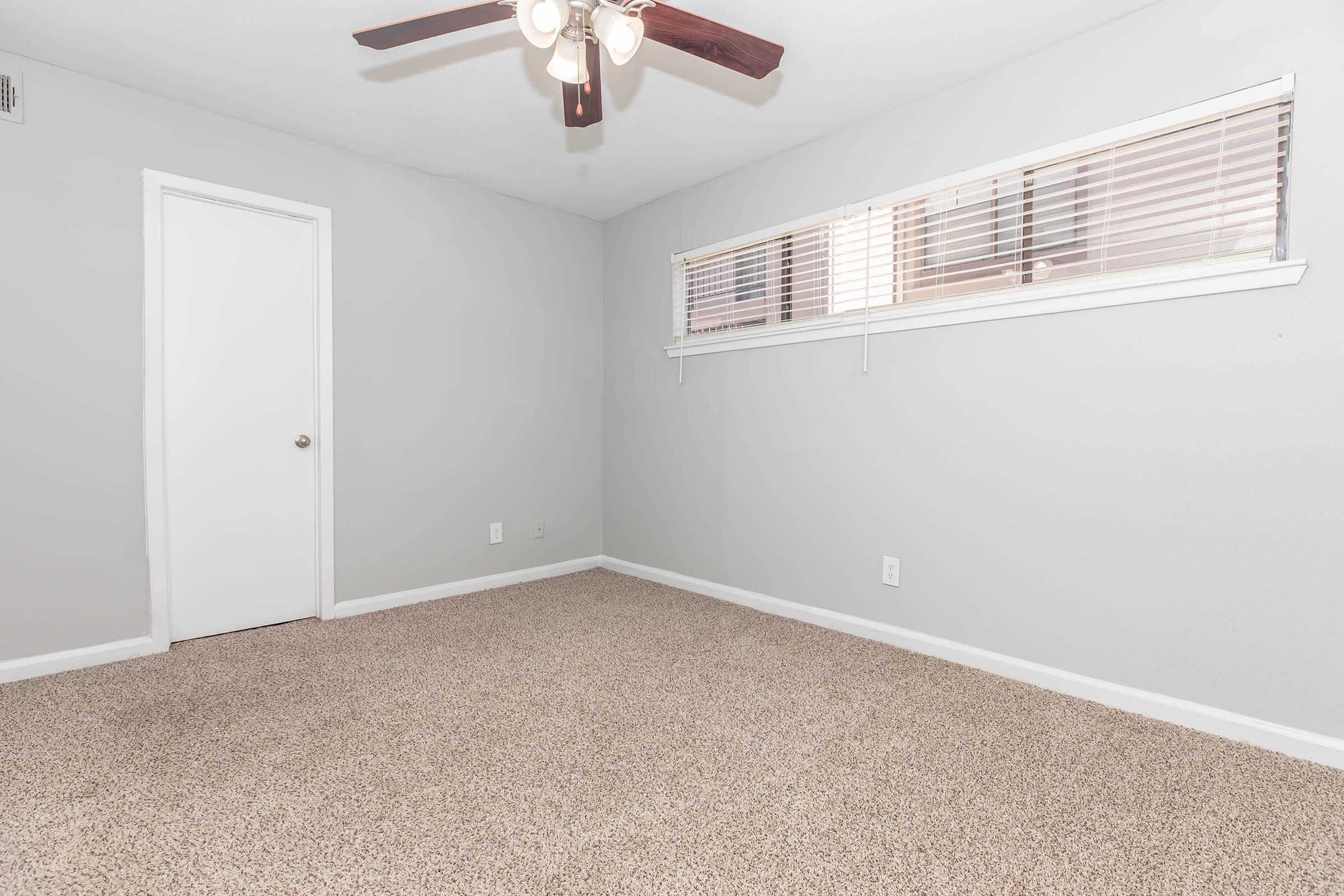 A vacant room with gray walls and a light brown carpet. There is a ceiling fan with wooden blades, a white door, and a large window with blinds. The space is well-lit and appears clean and minimalistic, suitable for various uses such as a bedroom or home office.
