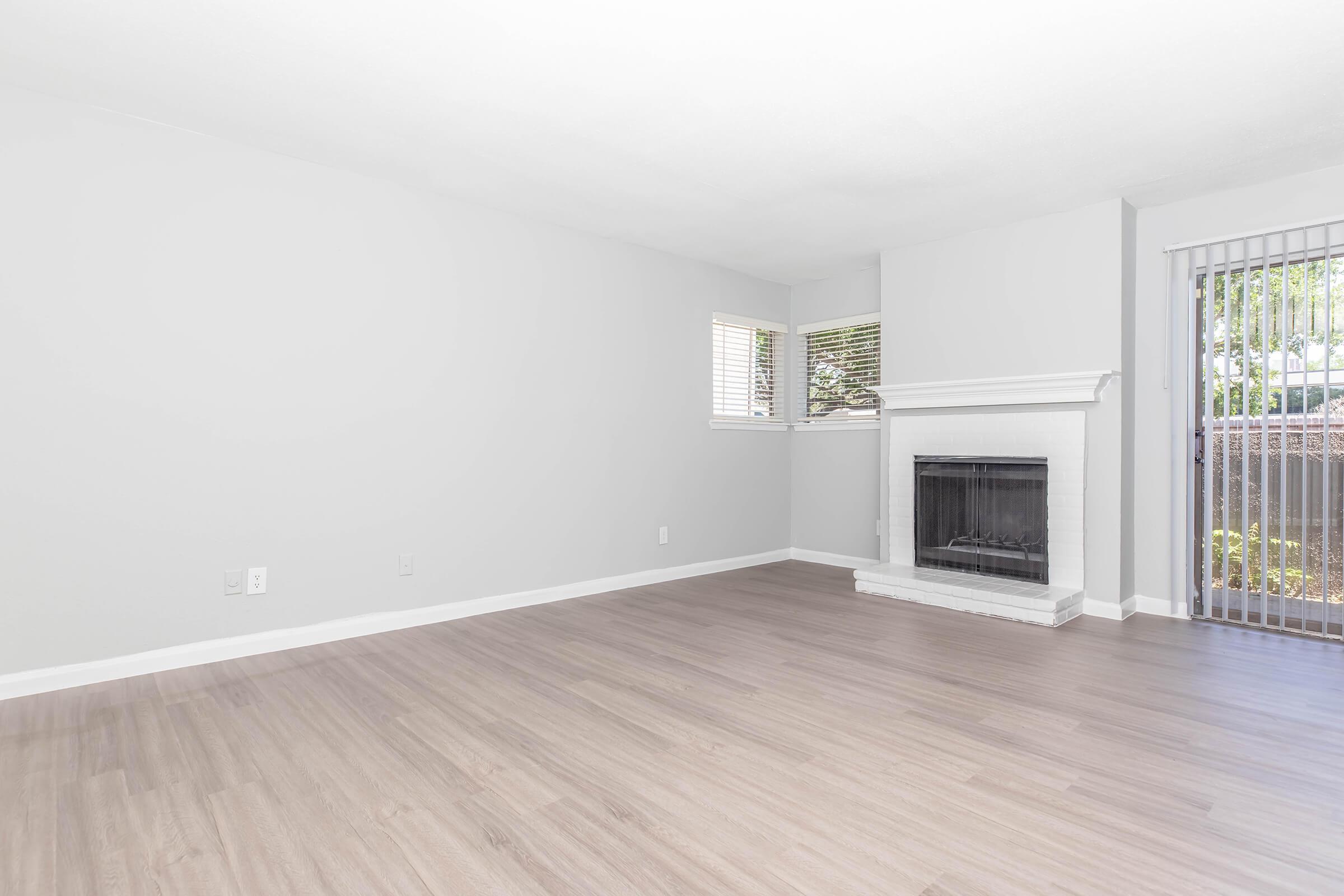 A spacious, empty living room with light gray walls and modern hardwood flooring. It features a white fireplace against one wall, a large window allowing natural light, and a sliding glass door leading outside. The overall ambiance is bright and inviting.
