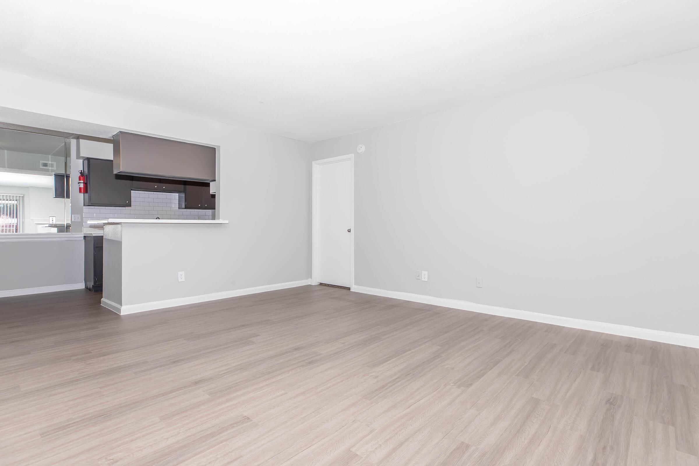 An empty living room with light gray walls and wooden-style flooring. There is a doorway leading to another room, and a partial wall with a bar counter that opens to a kitchen area in the background. The space is bright and airy, with no furniture or decorations.
