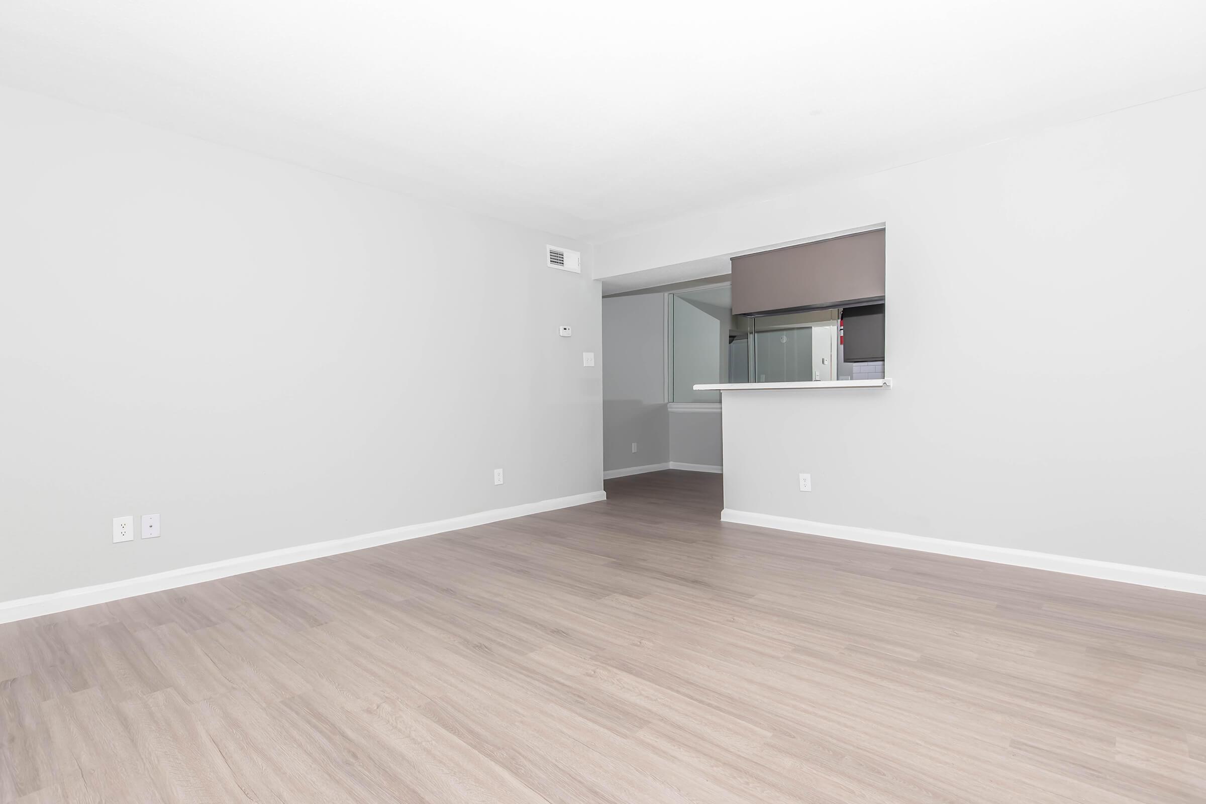 A spacious, empty room with light gray walls and a light wood floor. There's a small opening to an adjacent area, possibly a kitchen, indicated by a counter. The room is well-lit, giving a clean and modern feel.