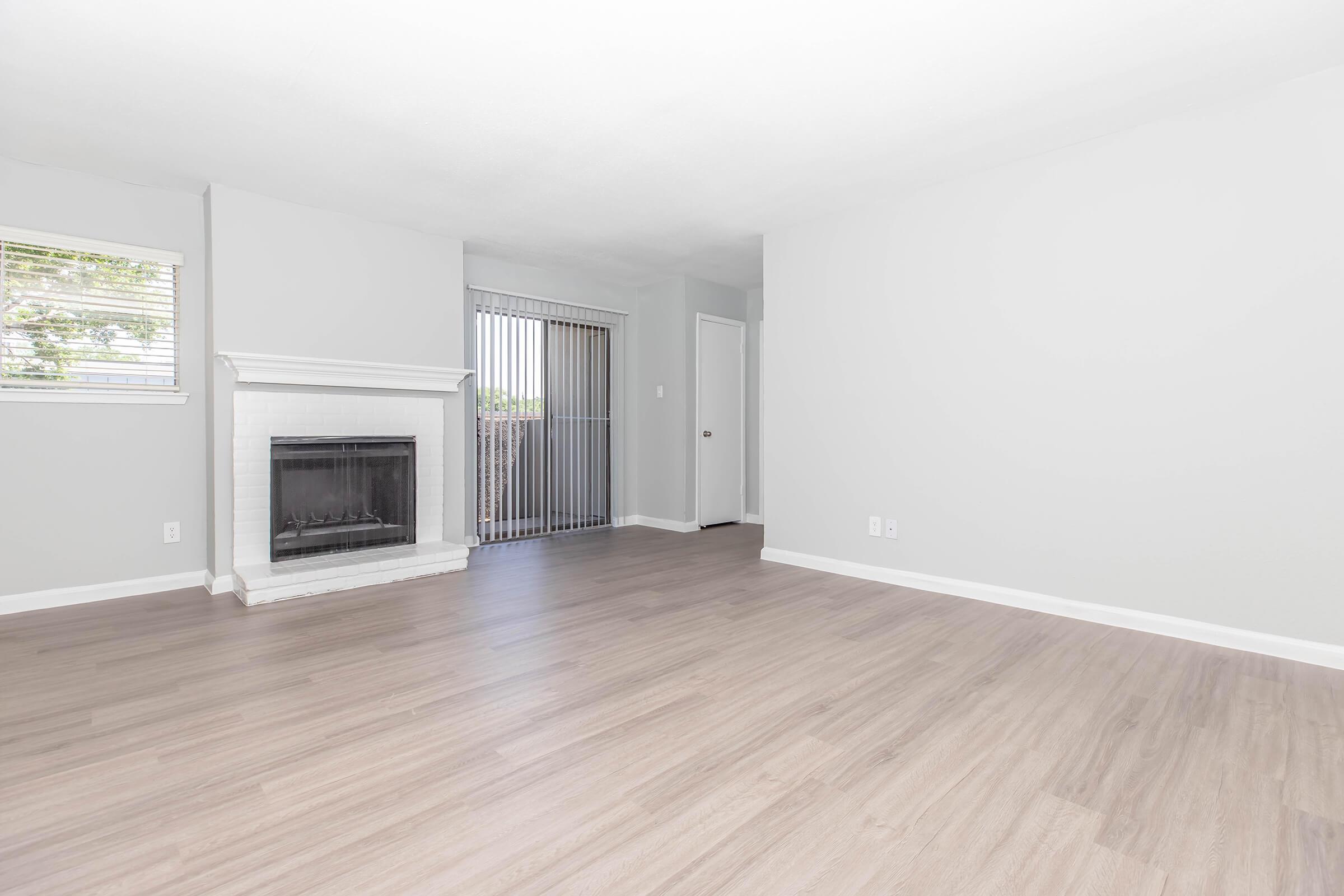 A spacious, empty living room with light-colored walls and a hardwood floor. A white fireplace is situated on one wall, and there are vertical blinds covering a sliding glass door that leads outside. A closed door is visible in the background, adding to the openness of the room.