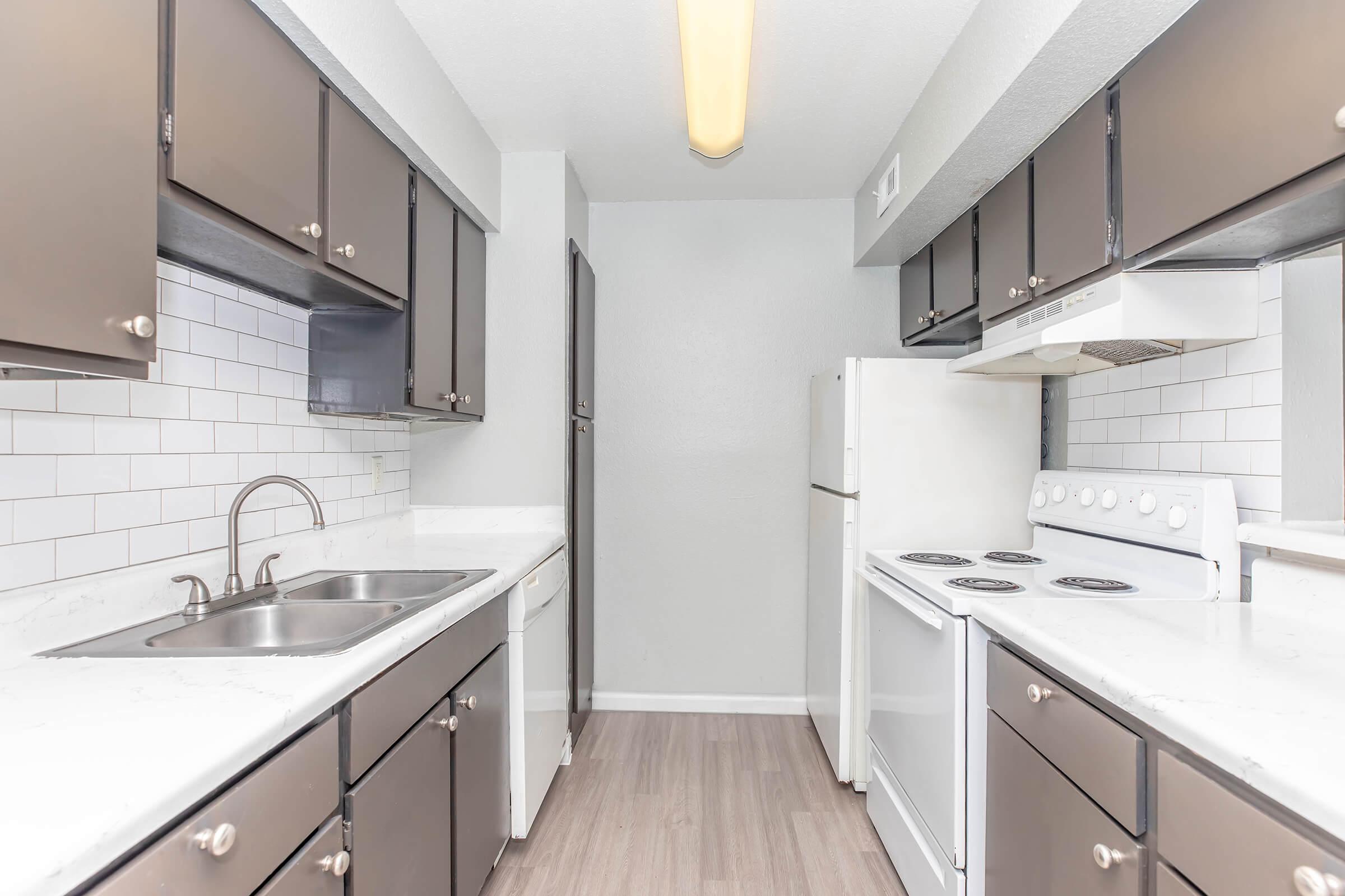 Modern kitchen featuring dark cabinets, a white tiled backsplash, stainless steel sink, and appliances including a stove and refrigerator. The space has a light-colored countertop and wood-style flooring, providing a clean and contemporary look. Well-lit with overhead lighting.