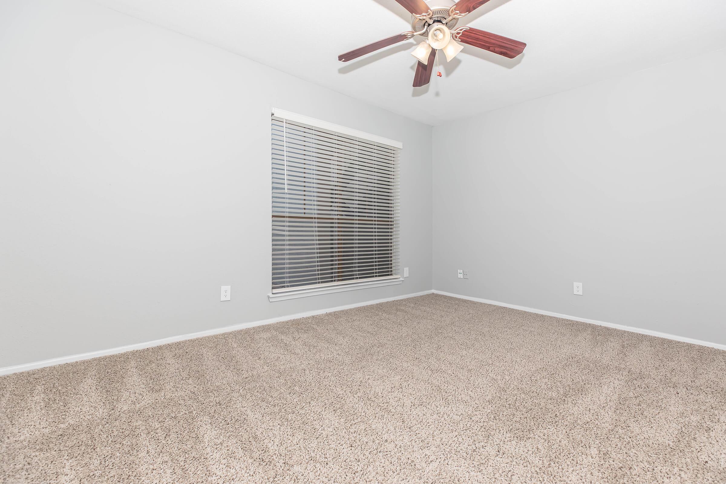 A vacant room with light gray walls and beige carpet. It features a ceiling fan with wooden blades and a window covered with horizontal blinds, allowing natural light to enter. The space is empty and unfurnished, highlighting the carpet texture and clean lines of the walls.
