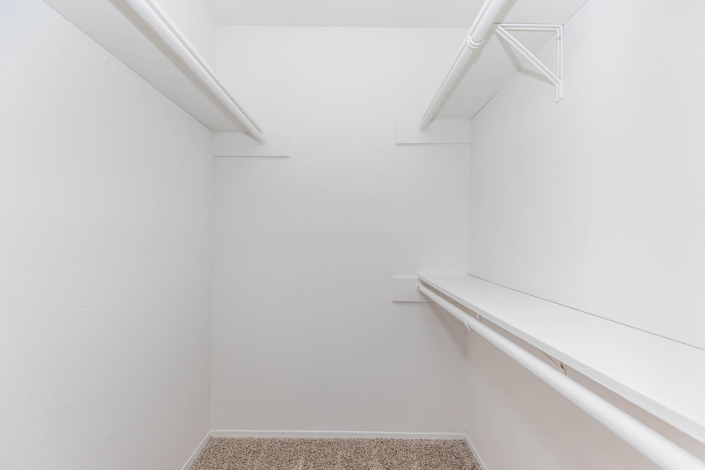 Empty closet with white walls and a carpeted floor. It features two hanging rods on opposite sides and a single shelf above, providing a minimalist storage space. Overall, the room appears clean and uncluttered, ready for organizational use.