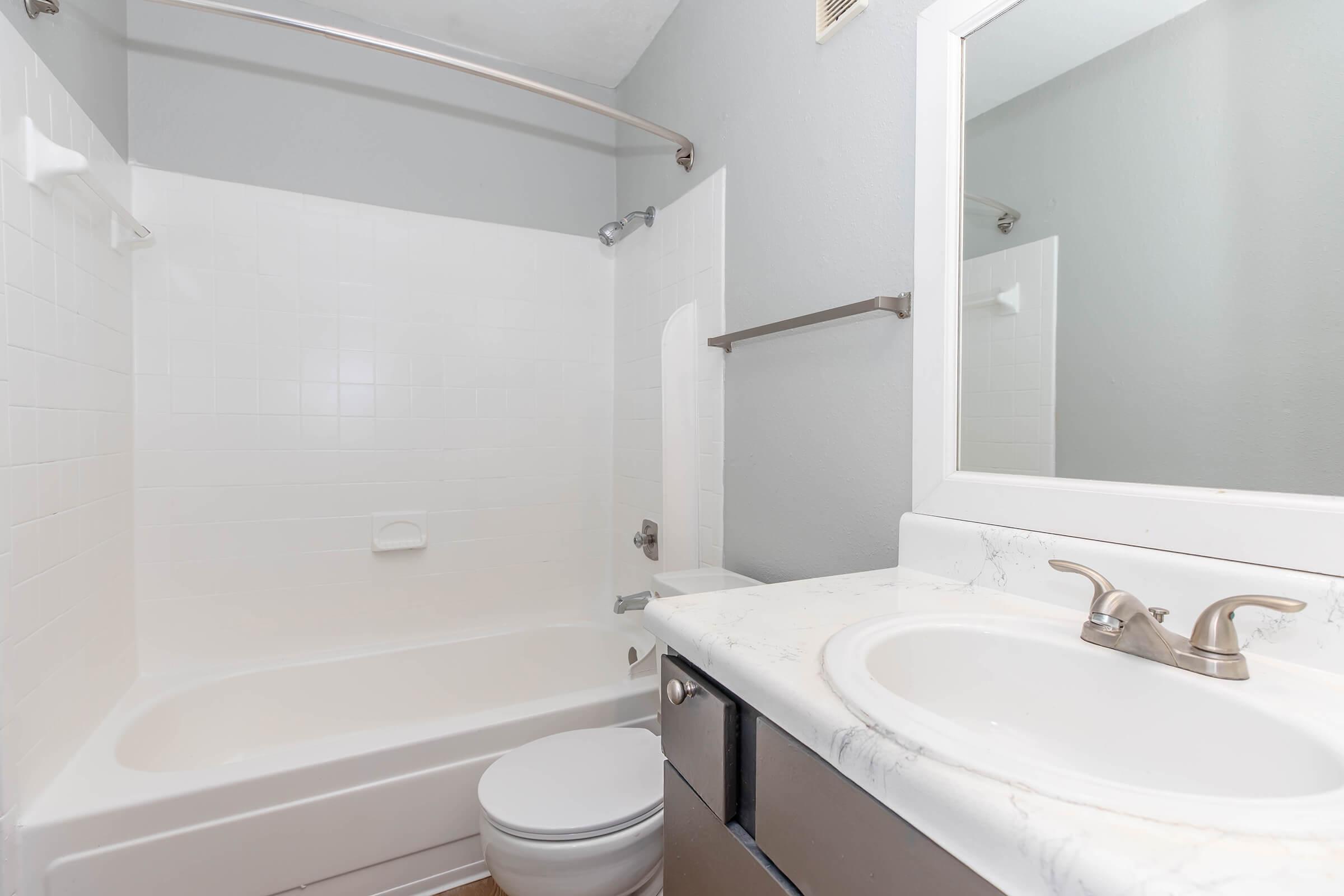 A clean and modern bathroom featuring a white bathtub with a shower, a toilet, and a vanity with a marble countertop and sink. The walls are painted a light gray, and there is a large mirror above the sink. The overall space is bright and minimalist.