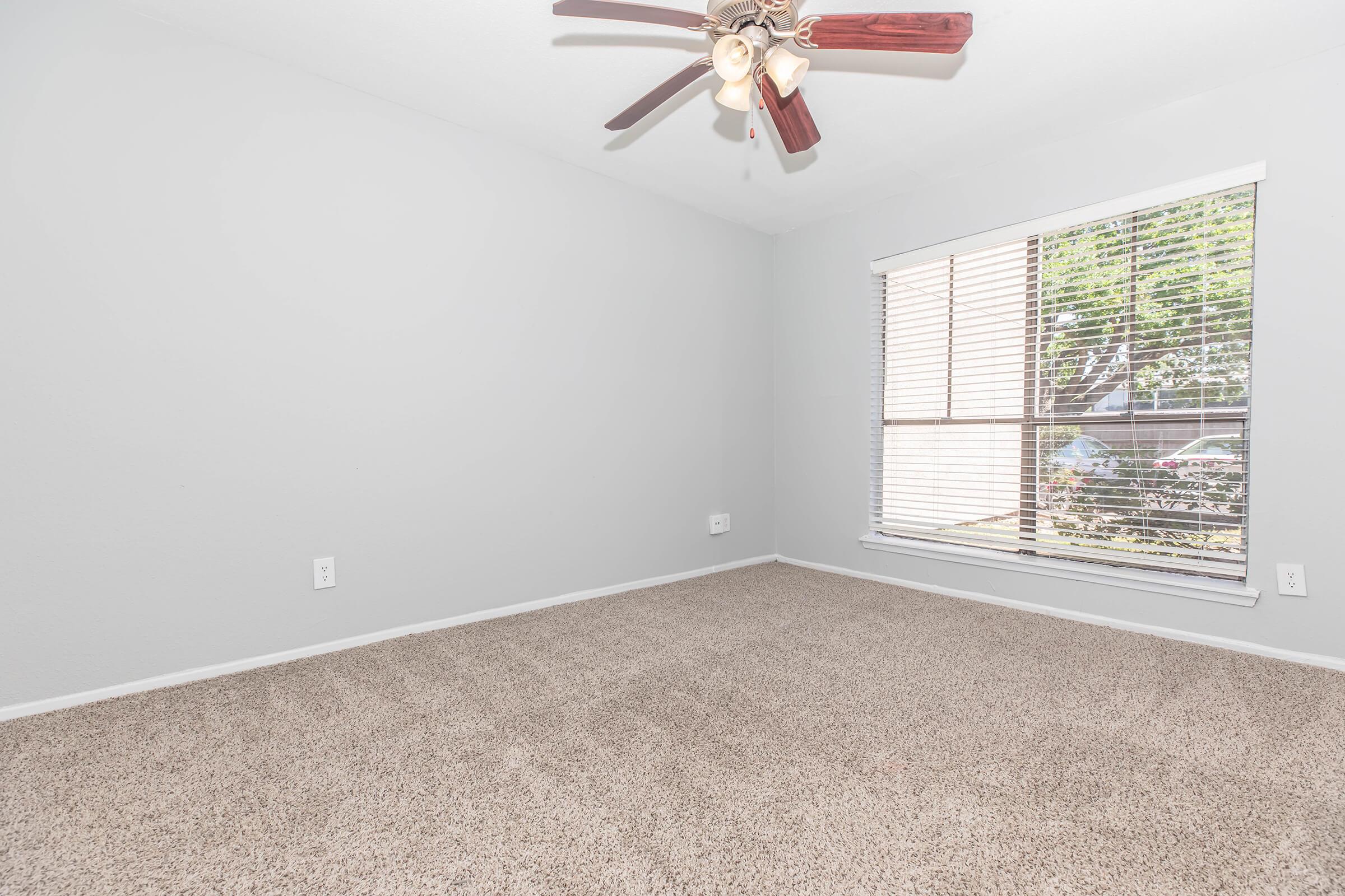 A well-lit, empty room featuring light gray walls and beige carpet. The space has a ceiling fan with wooden blades and a large window with blinds, allowing natural light to illuminate the room. The window offers a view of greenery outside.
