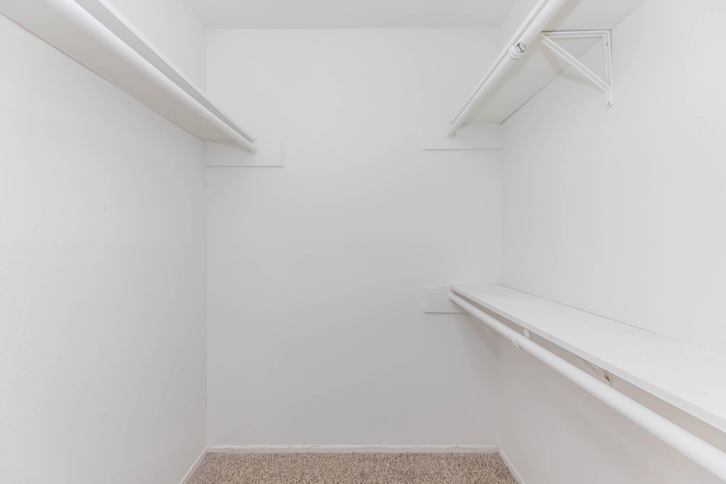 Empty closet with white walls and built-in shelves. The floor is carpeted, and there are two hanging rods on opposite walls, offering storage space for clothes or items. The overall appearance is clean and minimalist.