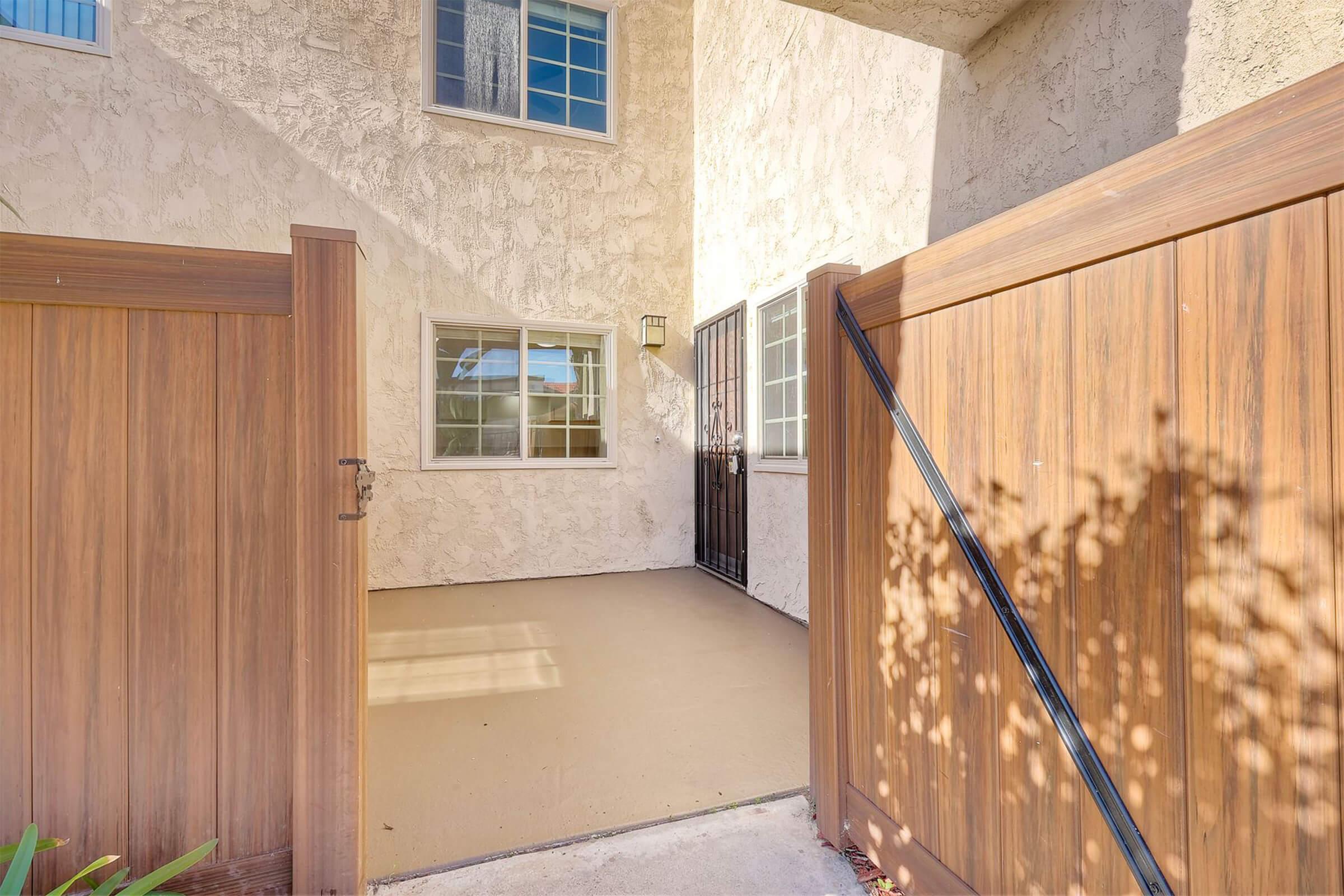 A view of an exterior entrance featuring a gated area with a wooden fence. The entrance has a smooth concrete surface and is framed by textured stucco walls, with windows on the side allowing natural light. The design is modern and clean, creating an inviting atmosphere.