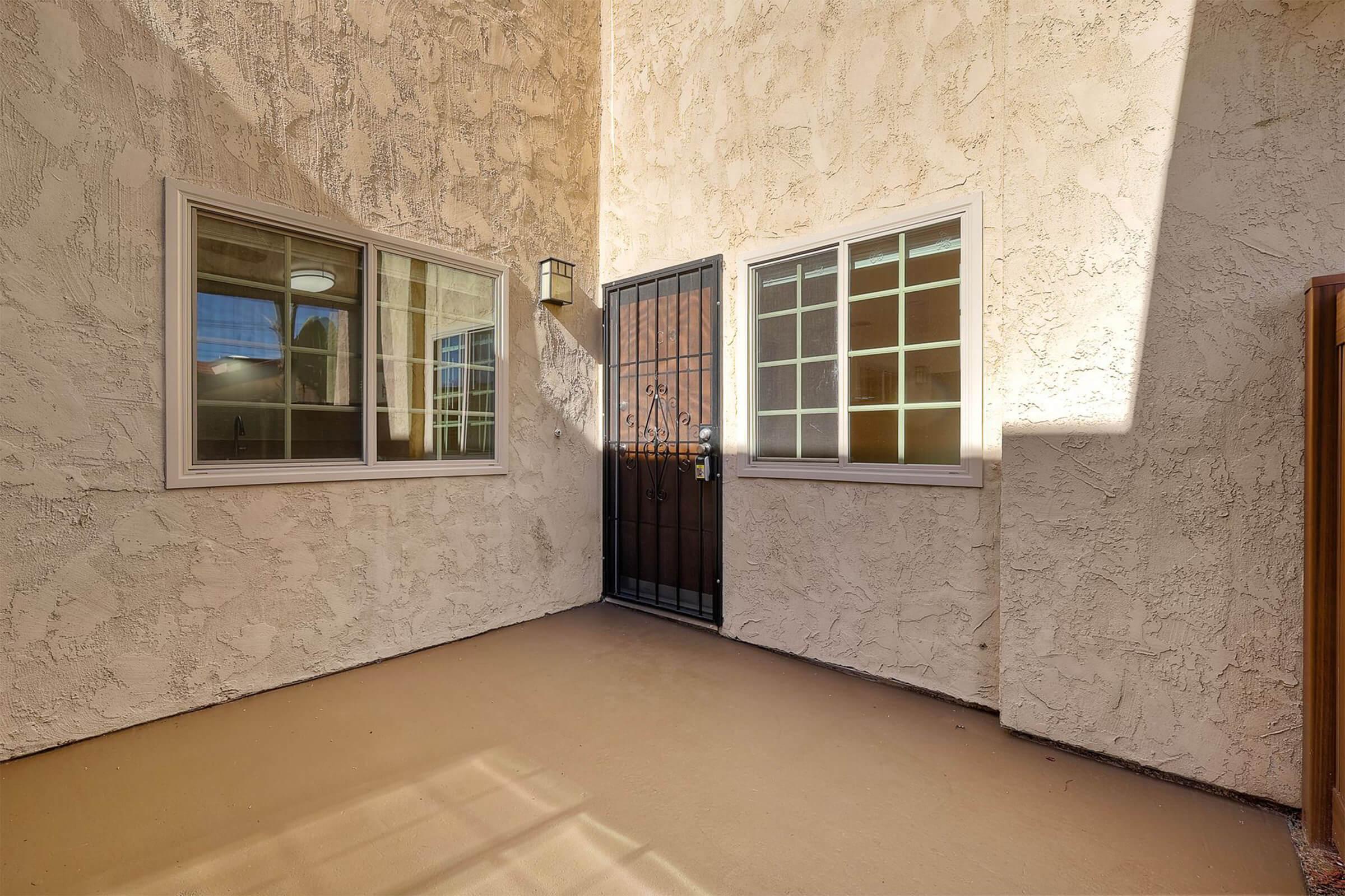 A small outdoor area featuring textured walls, a front door with a decorative metal grate, and two windows. The ground is finished with a smooth surface, and a light fixture is mounted beside the door, casting natural shadows in the sunlight.