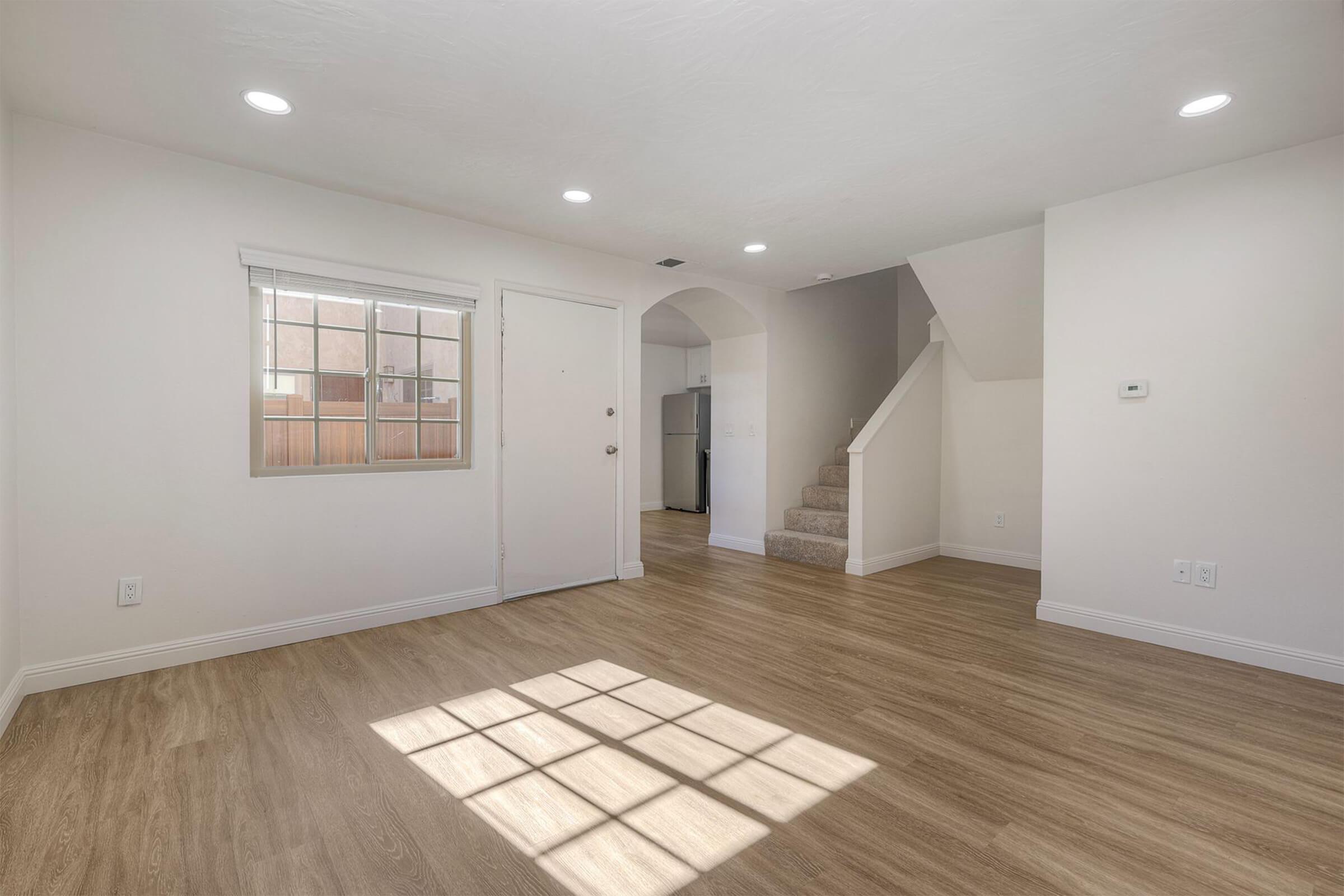 A bright, empty living room with wooden flooring, featuring a window with natural light, a front door, and access to a staircase. The walls are painted white, creating a clean and spacious feel, ideal for interior design or furnishing.