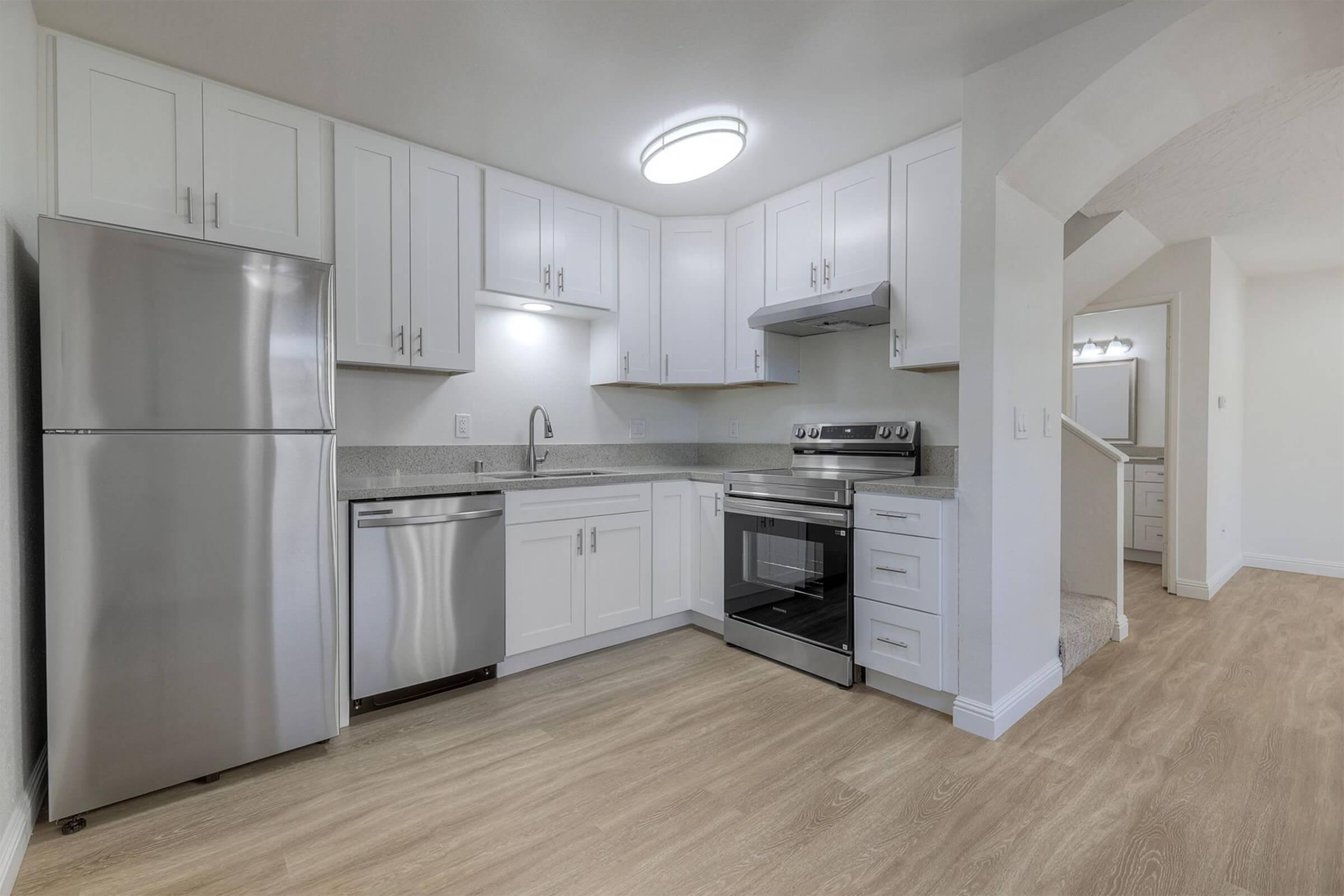 Modern kitchen featuring stainless steel appliances, including a refrigerator, dishwasher, and oven. White cabinetry and a light-colored countertop contrast with the warm wood vinyl flooring. Bright overhead lighting enhances the spacious, clean design. The kitchen is part of an open layout with a staircase in the background.