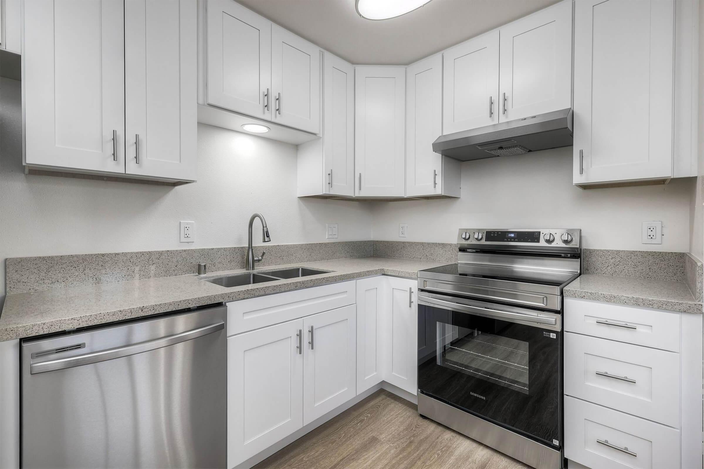 Modern kitchen featuring white cabinetry, a stainless steel sink, and a sleek stainless steel refrigerator. The kitchen includes a modern oven with a range hood, and gray countertops. Bright, well-lit space with ample storage and minimalist design.