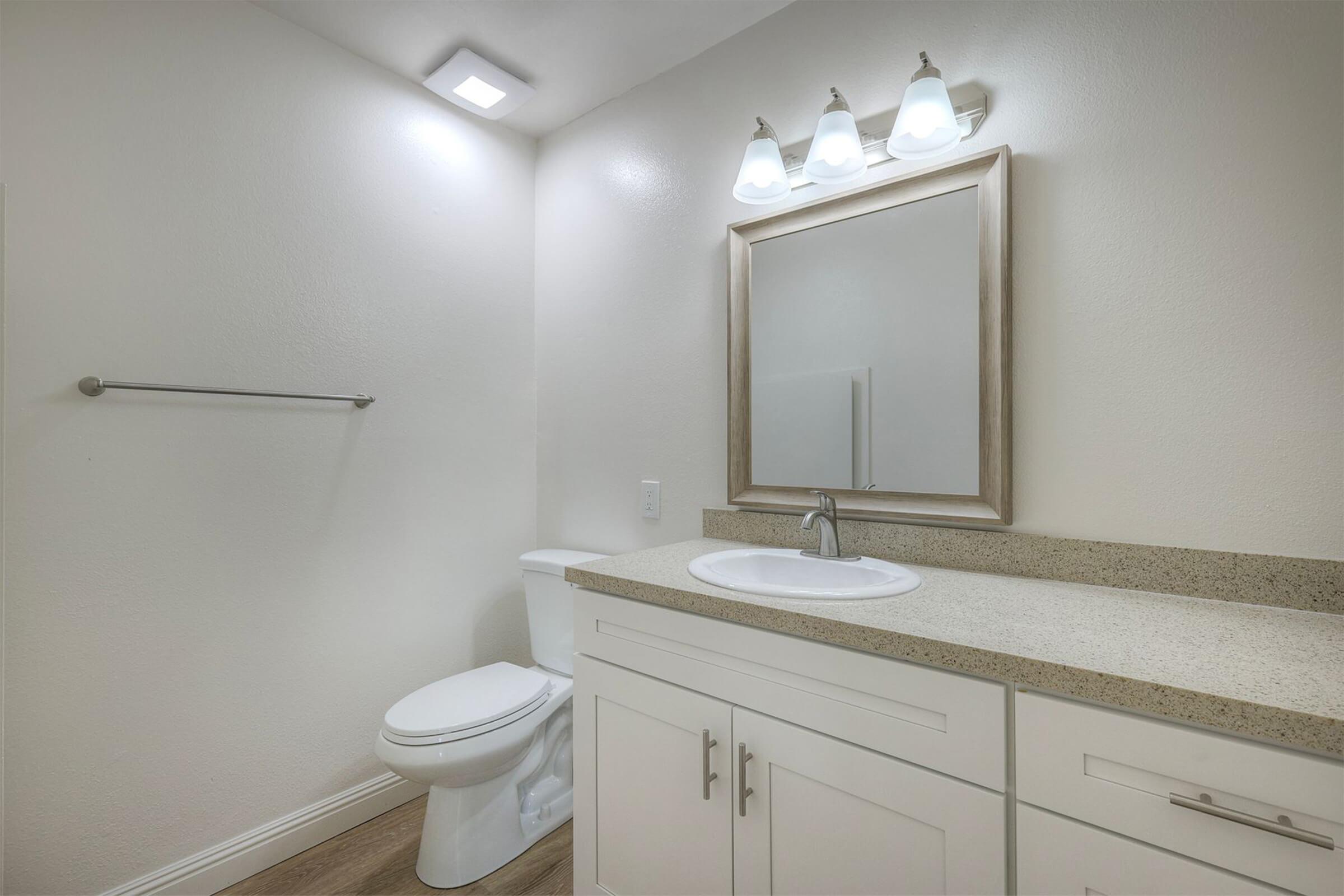 A clean and modern bathroom featuring a white toilet, a countertop sink with a silver faucet, and a wall mirror framed in wood. The walls are painted in a light color, and there are three light fixtures above the mirror. A towel rack is mounted on the wall, and the flooring has a wood-like appearance.
