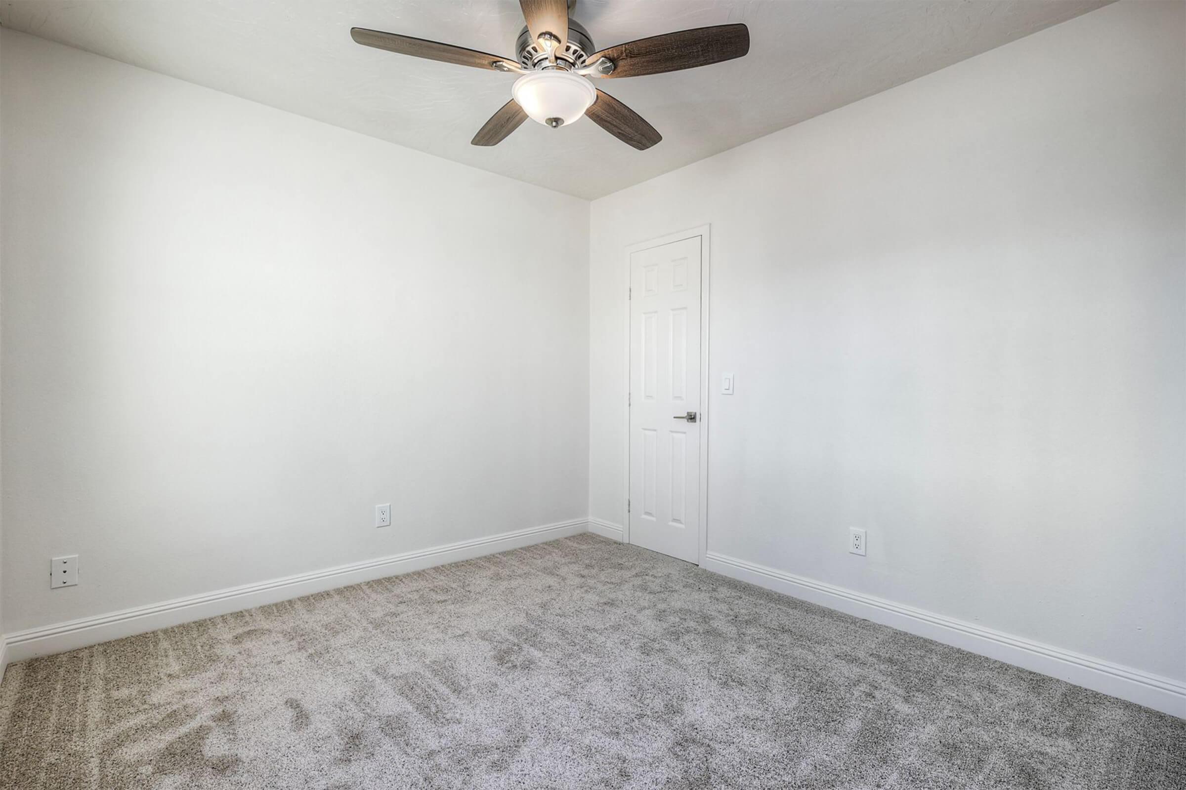 A clean, empty room with light gray carpet and white walls. Ceiling fan with wooden blades is mounted on the ceiling. A white door is visible, leading to another area. The room is well-lit, showcasing its spaciousness and simplicity.