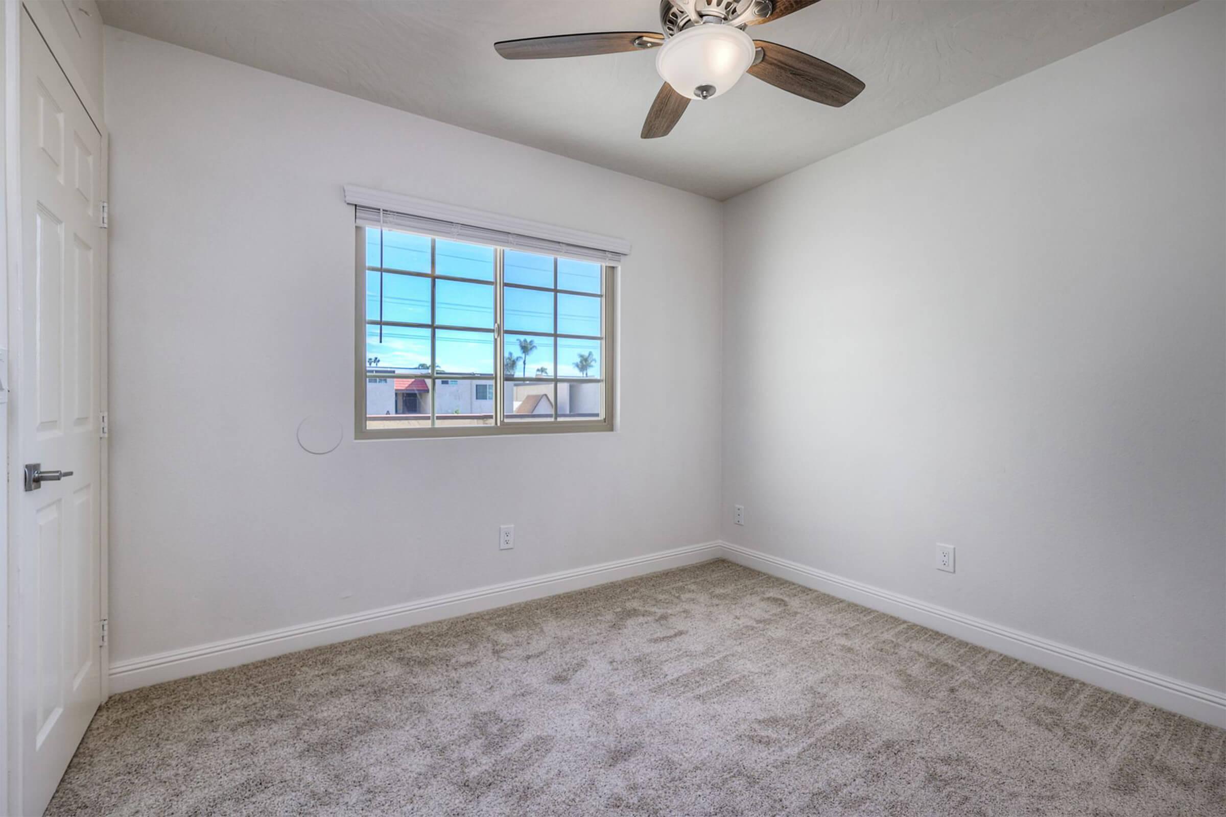 Empty room featuring light-colored walls, a ceiling fan, and carpeted flooring. A window allows natural light to enter, revealing a clear sky and distant landscape. The room has a simple, modern design with a door visible on the left side.