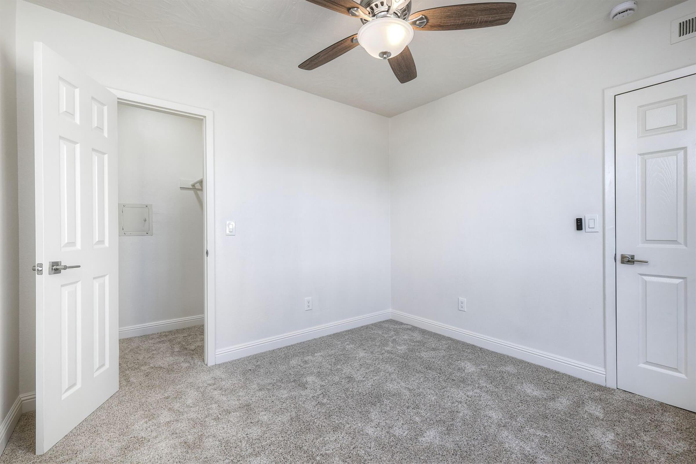 A well-lit, empty room featuring beige carpet flooring, white walls, and a ceiling fan. There are two doors: one leading to a closet and another to an exterior area. The room is spacious with minimal decoration, creating a clean and neutral atmosphere.