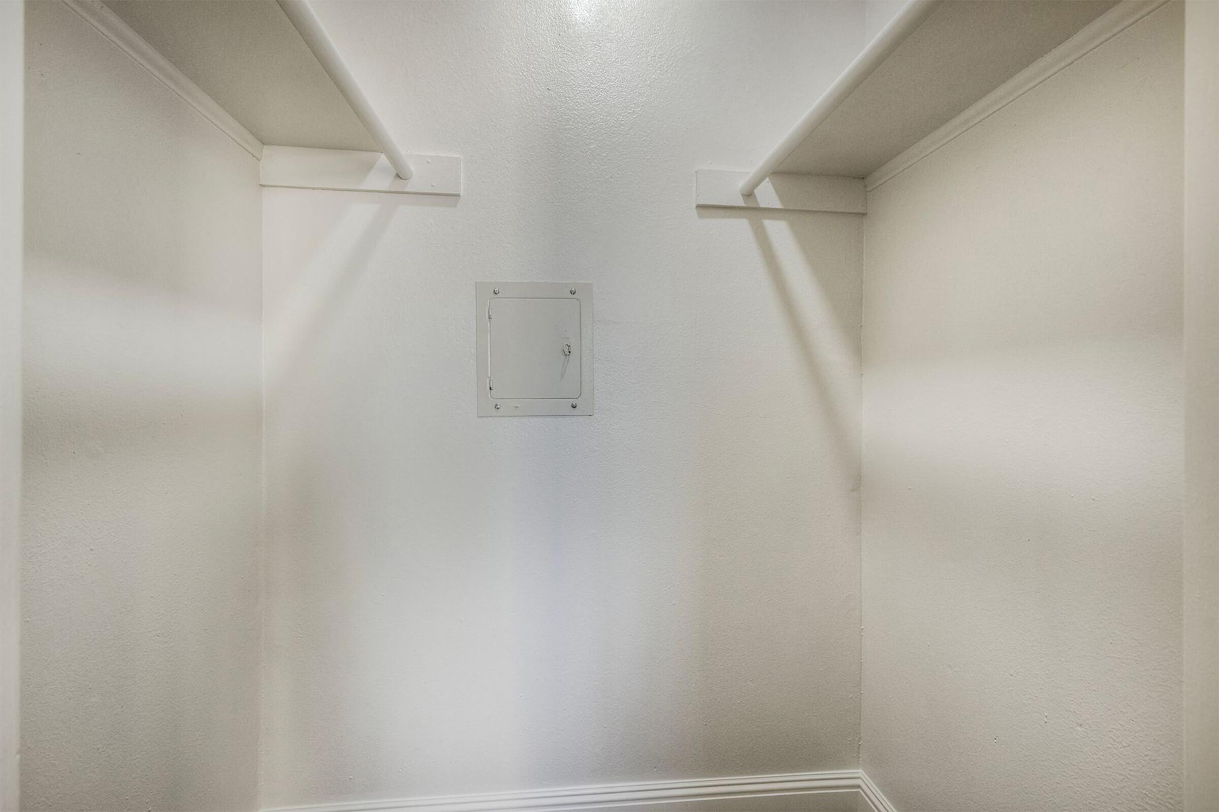 Empty closet with white walls, a small panel on the back wall, and two shelves on either side. The lighting is bright, highlighting the spaciousness of the area.