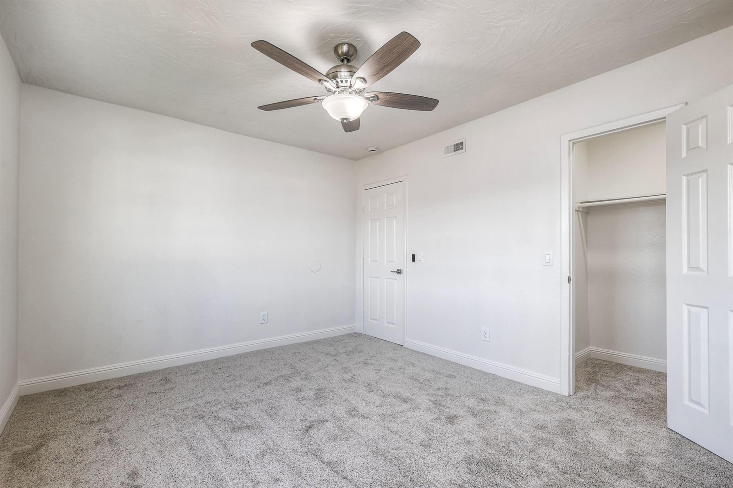Empty room with light-colored walls and a ceiling fan. The floor is covered in light gray carpet. A closet door is partially visible on the right. The room has a bright atmosphere with ample natural light.