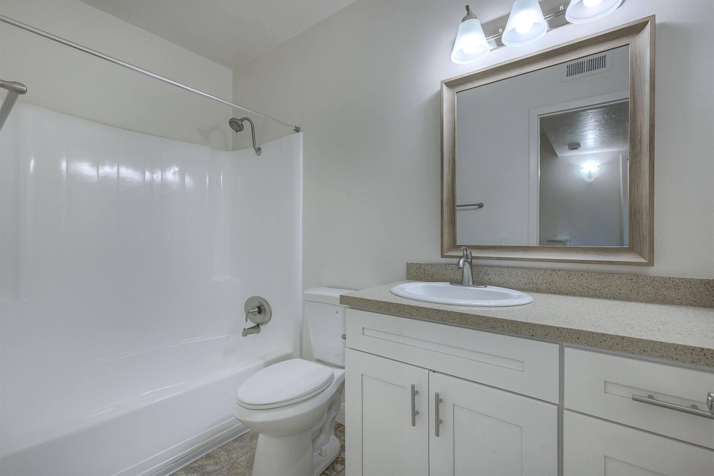 A clean, modern bathroom featuring a white bathtub with a glass shower door, a white toilet, and a vanity with a sink. The walls are painted in a light color, and there is a large mirror above the sink. Bright lighting enhances the spacious feel of the room.