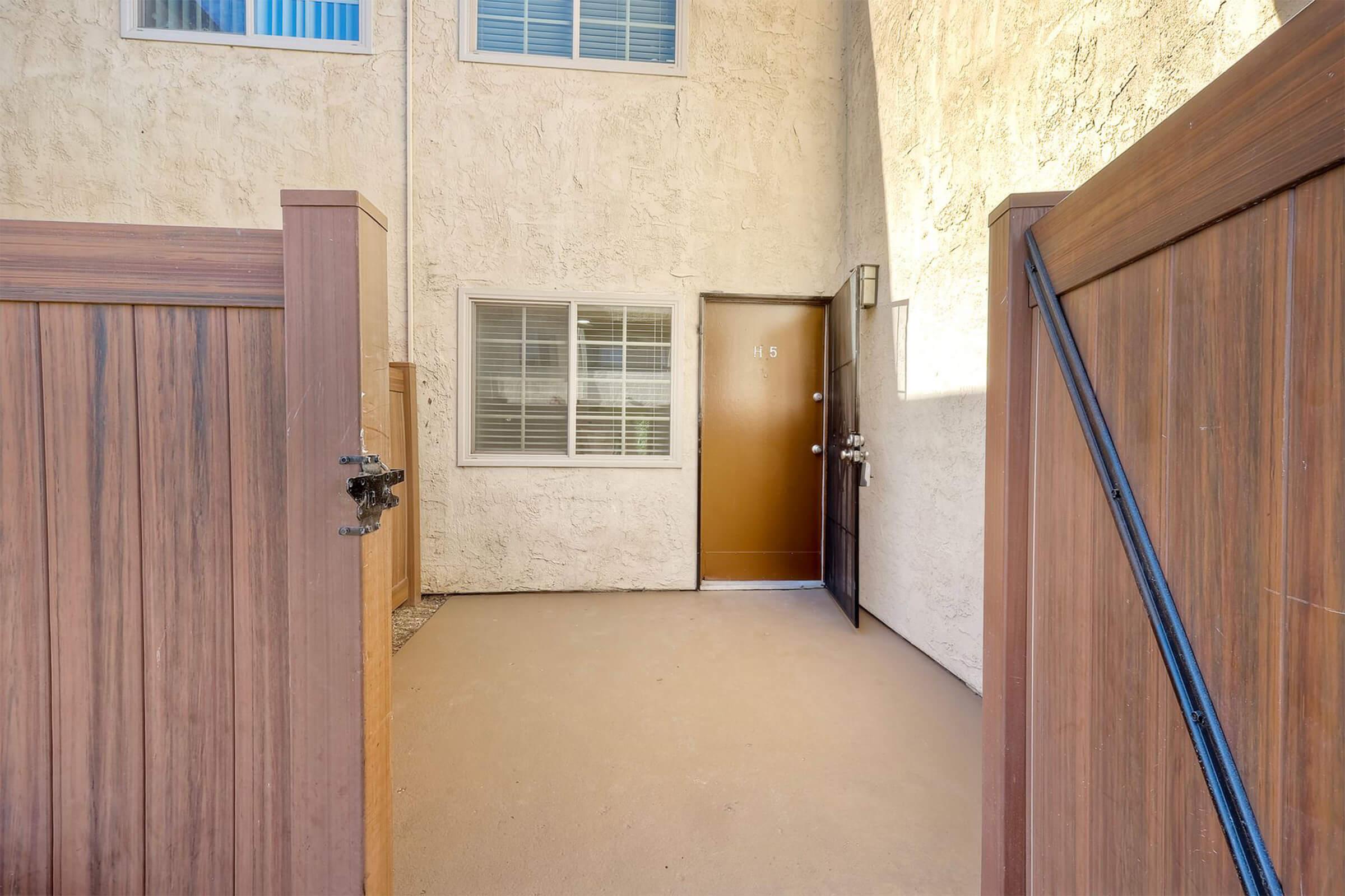 A small entryway with a brown door set into a textured beige wall. The area is enclosed by wooden gates on either side, leading to a patio or outdoor space. A window is visible to the left of the door, allowing natural light into the entrance. The overall space appears clean and well-maintained.