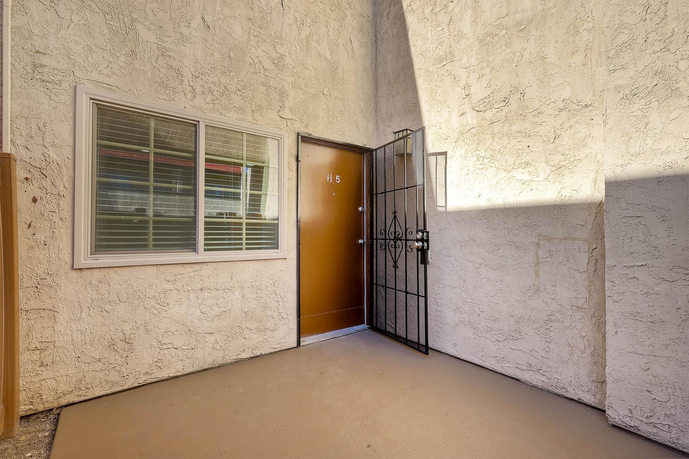 A view of a beige exterior wall with a window beside a brown door, which has a security gate. Sunlight casts shadows on the textured surface. The ground is flat and unadorned, creating a simple entryway. The door is labeled "H5."