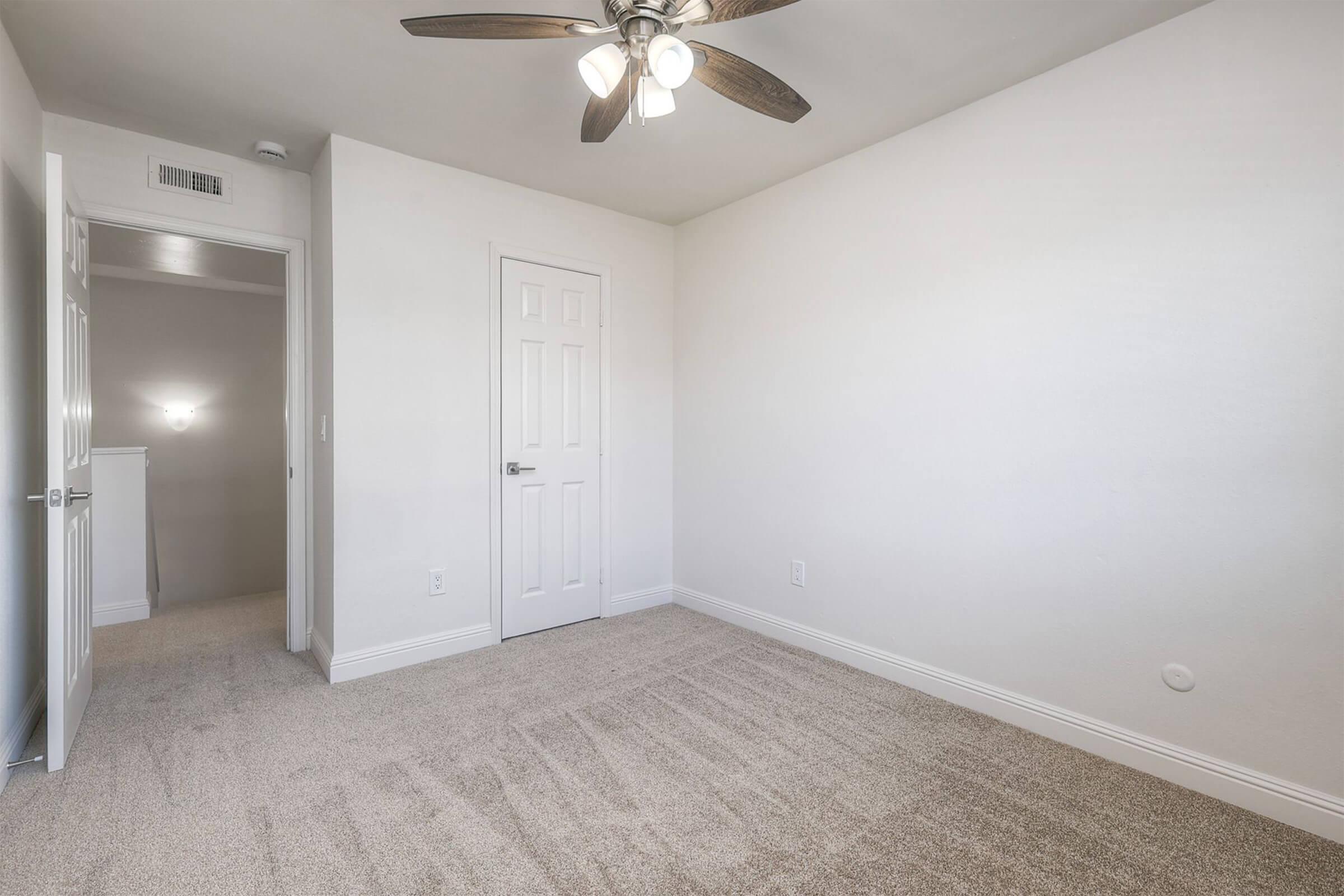 Empty room with light beige carpet, white walls, and a ceiling fan with three bulbs. The room features a closed door with a knob and a small hallway leading out of the space. A single light source illuminates the room, creating a bright and open atmosphere.