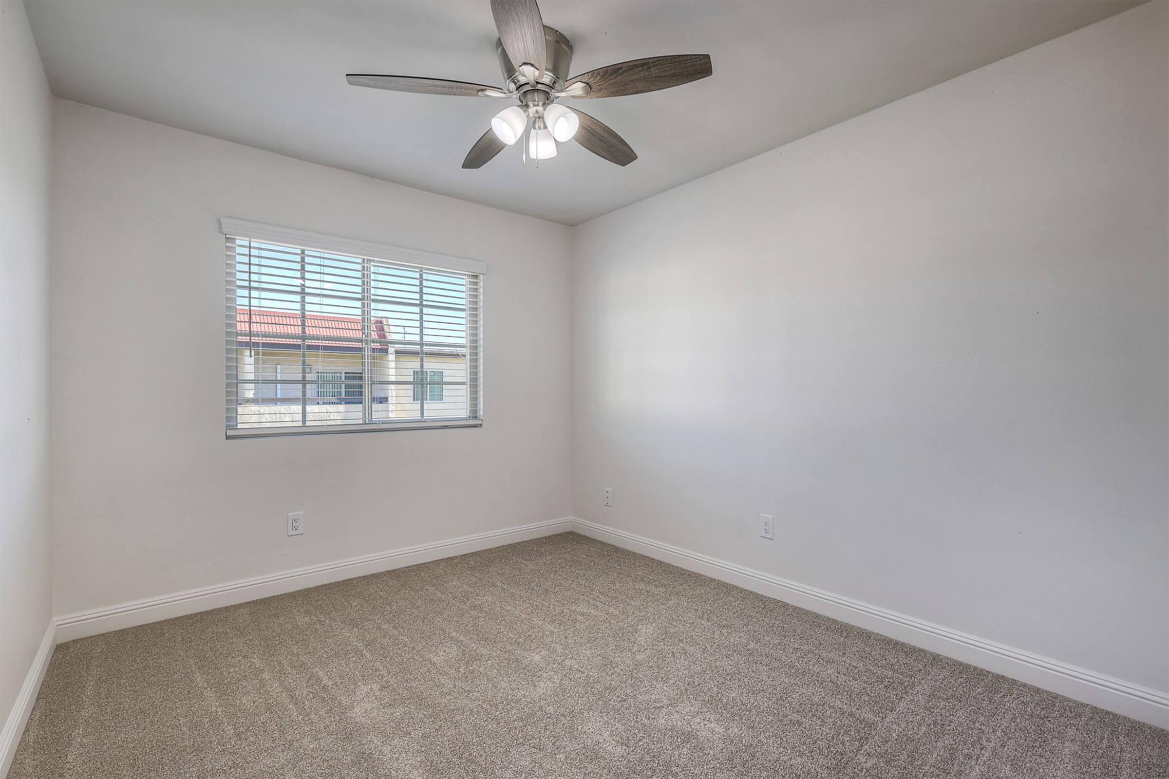 Empty room with light gray carpet, a ceiling fan with four light bulbs, and a window featuring white blinds. The walls are painted a soft beige, and there's no furniture present, creating a spacious ambiance. Natural light enters through the window, illuminating the room.