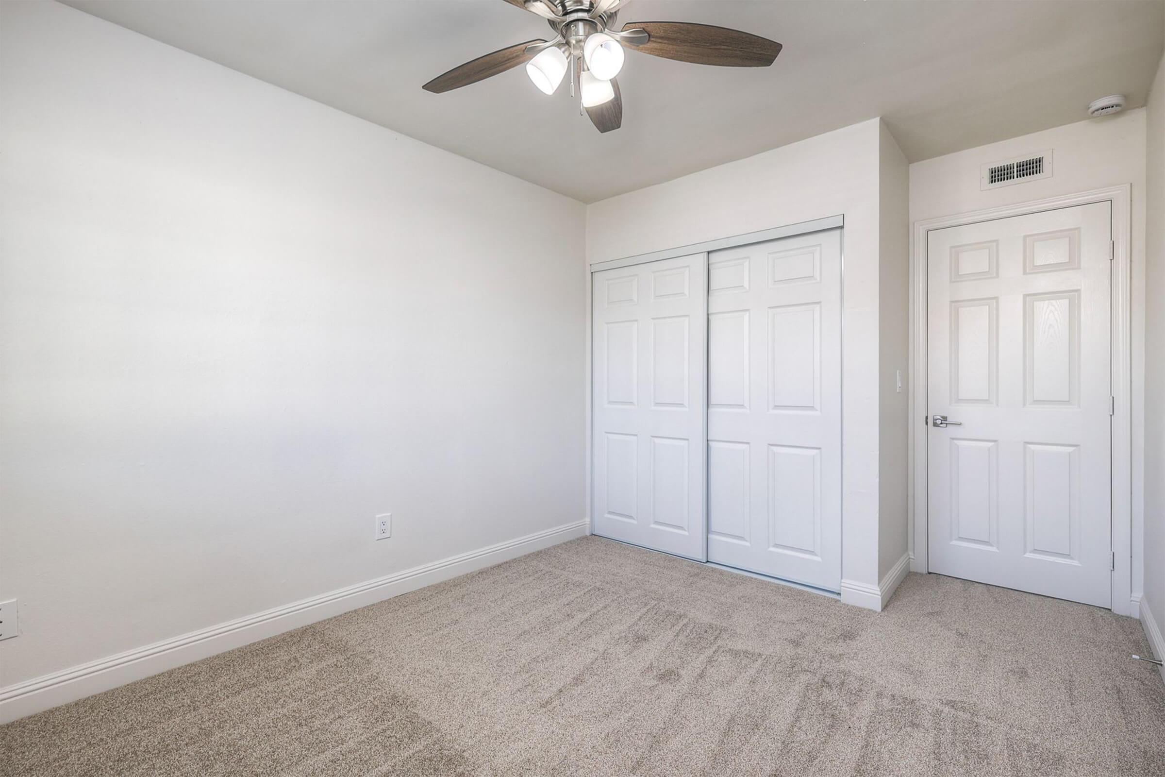 A spacious, empty room with light beige carpet, white walls, and a ceiling fan. The room features a double-door closet and a white door leading outside. Natural light brightens the space, giving it a clean and fresh appearance.