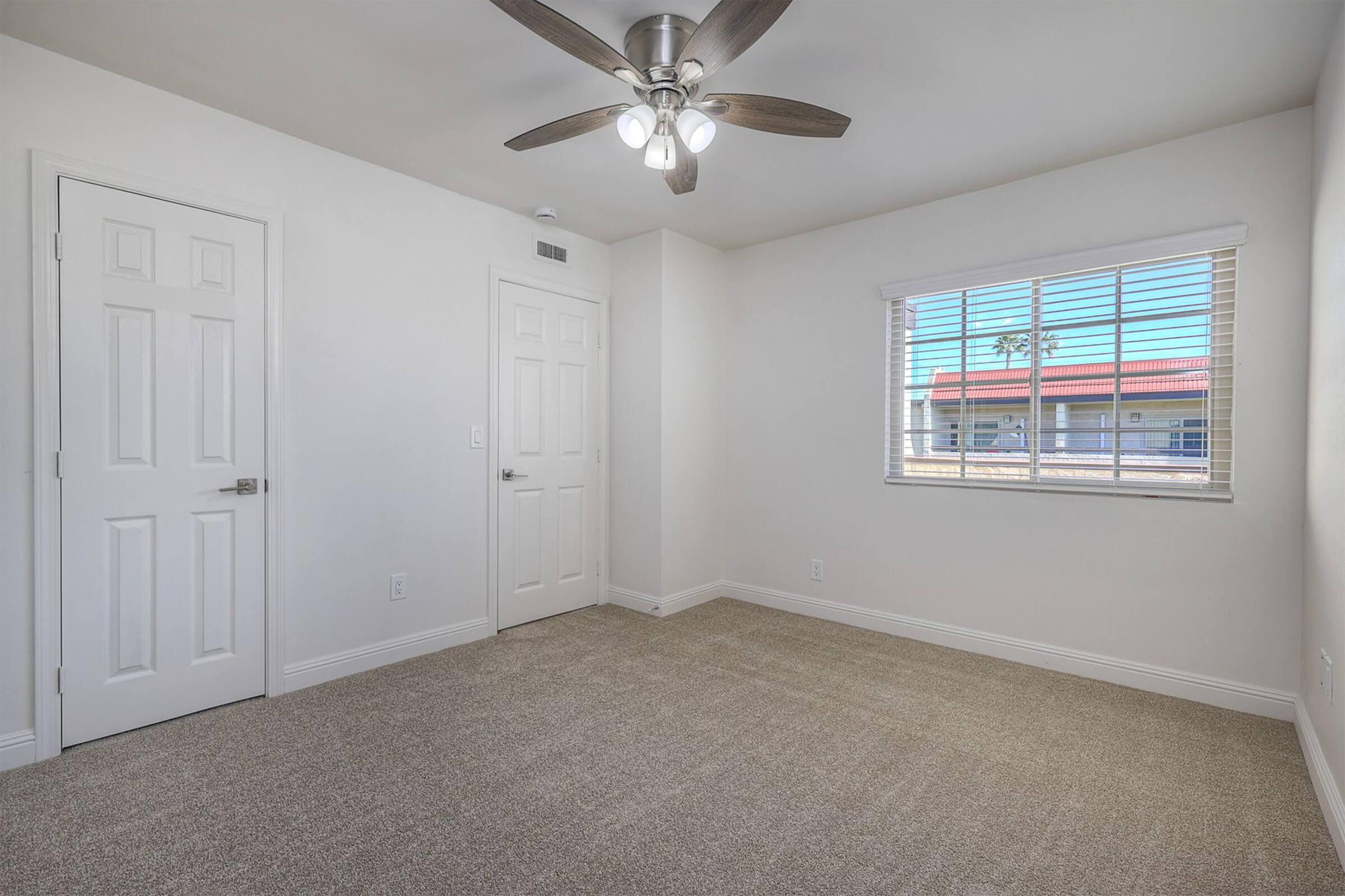 A spacious, empty room with light beige carpet and white walls. A ceiling fan with wooden blades is mounted above. Natural light enters through a window with horizontal blinds, revealing a view of a red building outside. Two white doors are positioned on the left side of the room.