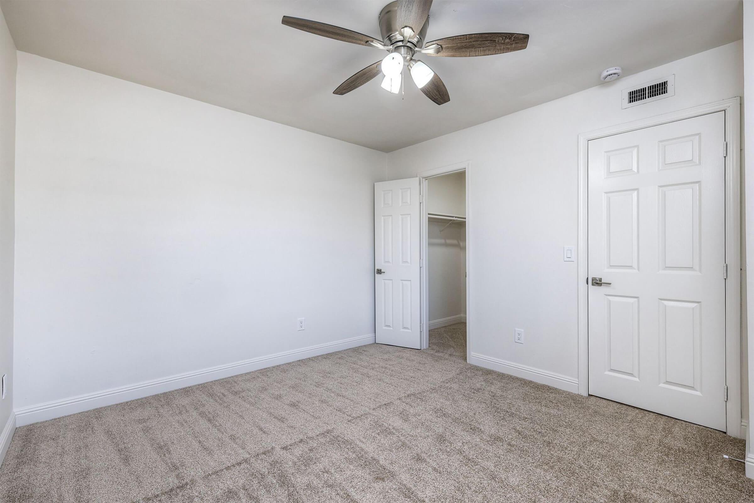 Empty room with light-colored walls and carpet. A ceiling fan with wooden blades hangs from the ceiling. An open door leads to a closet on the left side, with a closed door on the right side. The room has a minimalist aesthetic, emphasizing space and brightness.